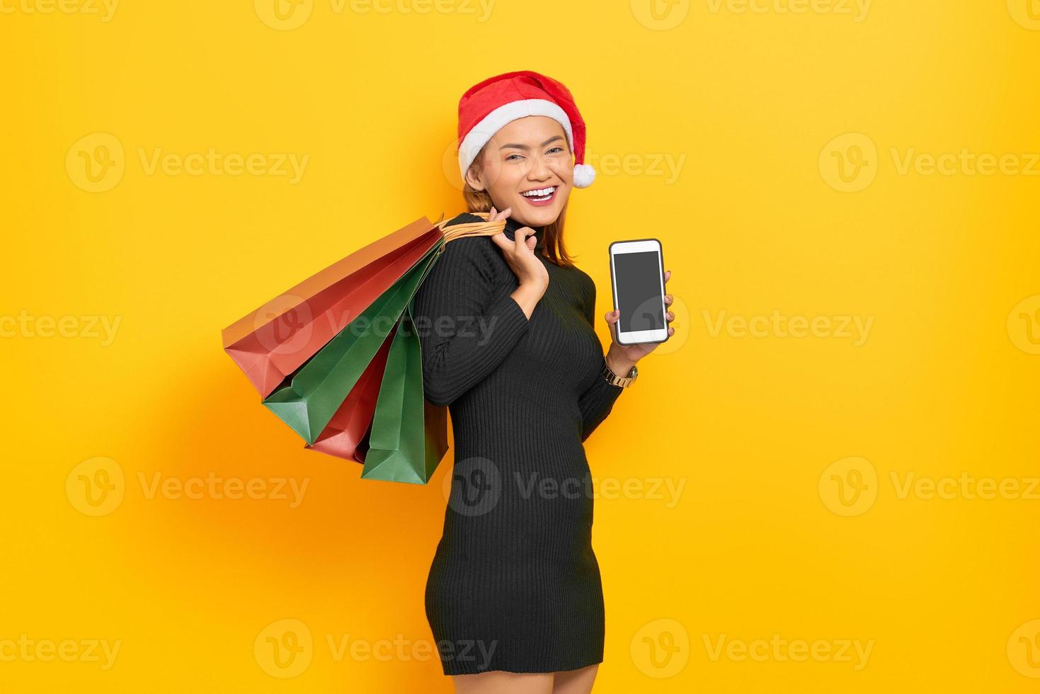 Sonriente joven mujer asiática con sombrero de santa claus mostrando teléfono móvil con pantalla en blanco y sosteniendo bolsas de la compra aisladas sobre fondo amarillo foto