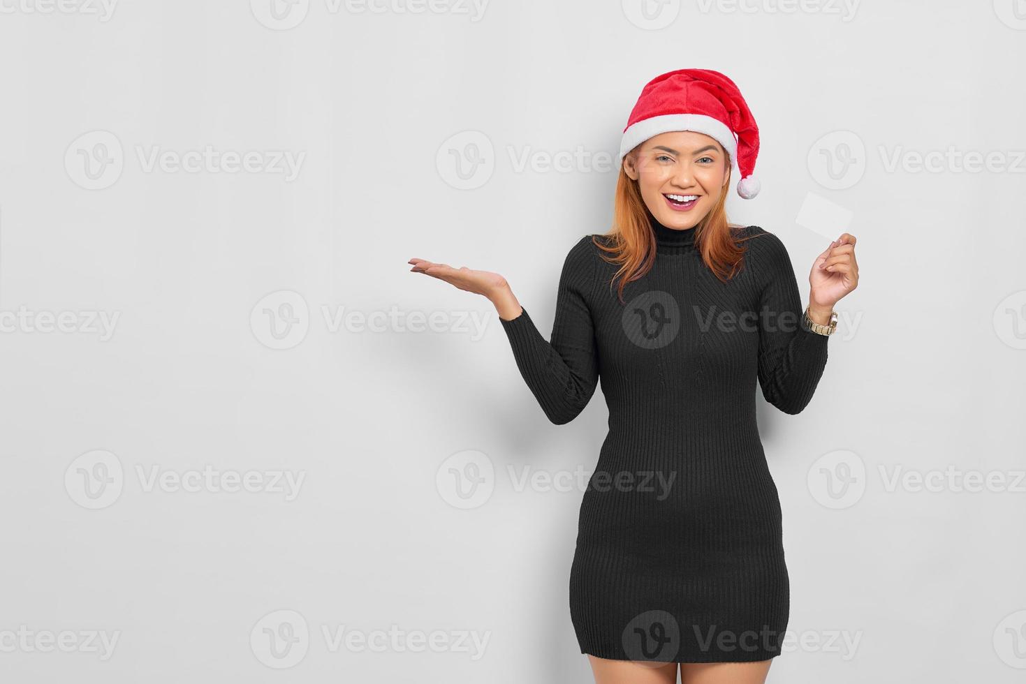 Sonriente joven mujer asiática con sombrero de santa claus presentando espacio de copia y sosteniendo una tarjeta en blanco aislada sobre fondo blanco. foto