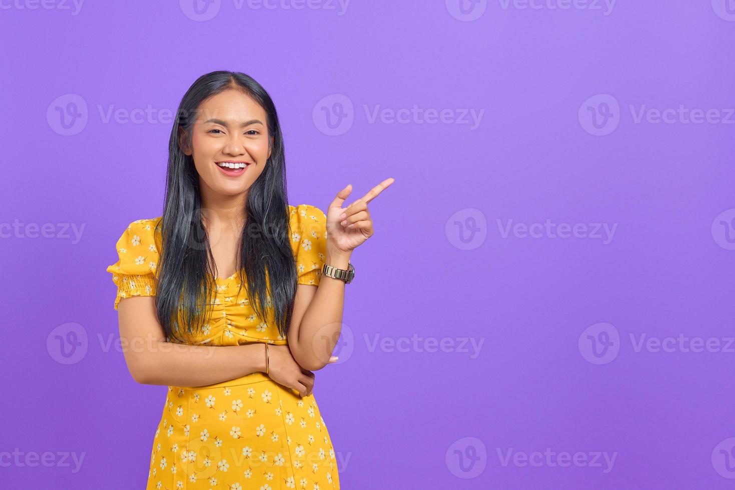 Portrait of smiling young Asian woman pointing finger at copy space on purple background photo