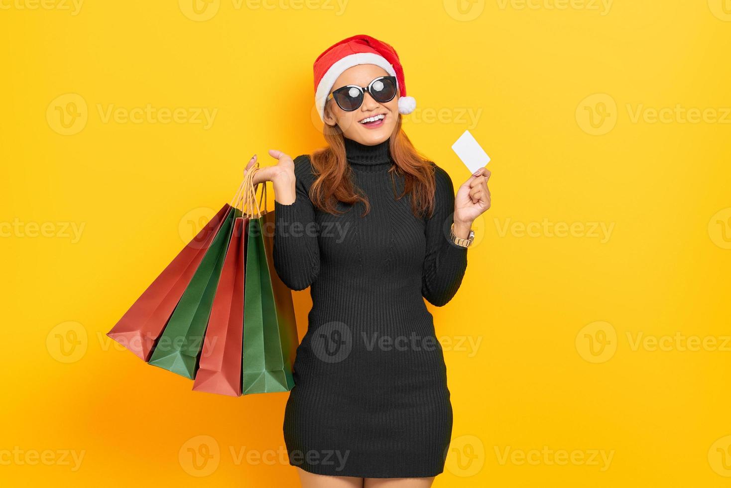 Sonriente joven mujer asiática con gorro de Papá Noel y gafas de sol sosteniendo bolsas de la compra y tarjeta de plástico aislado sobre fondo amarillo foto