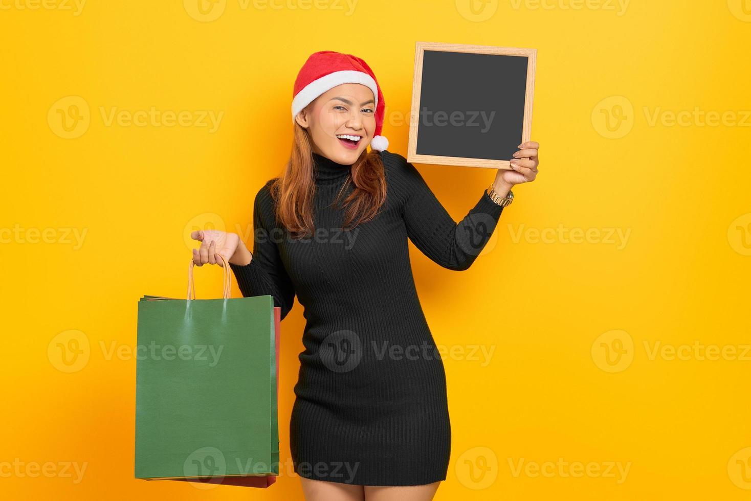 Smiling young Asian woman in Santa Claus hat holding shopping bags and chalkboard isolated over yellow background photo