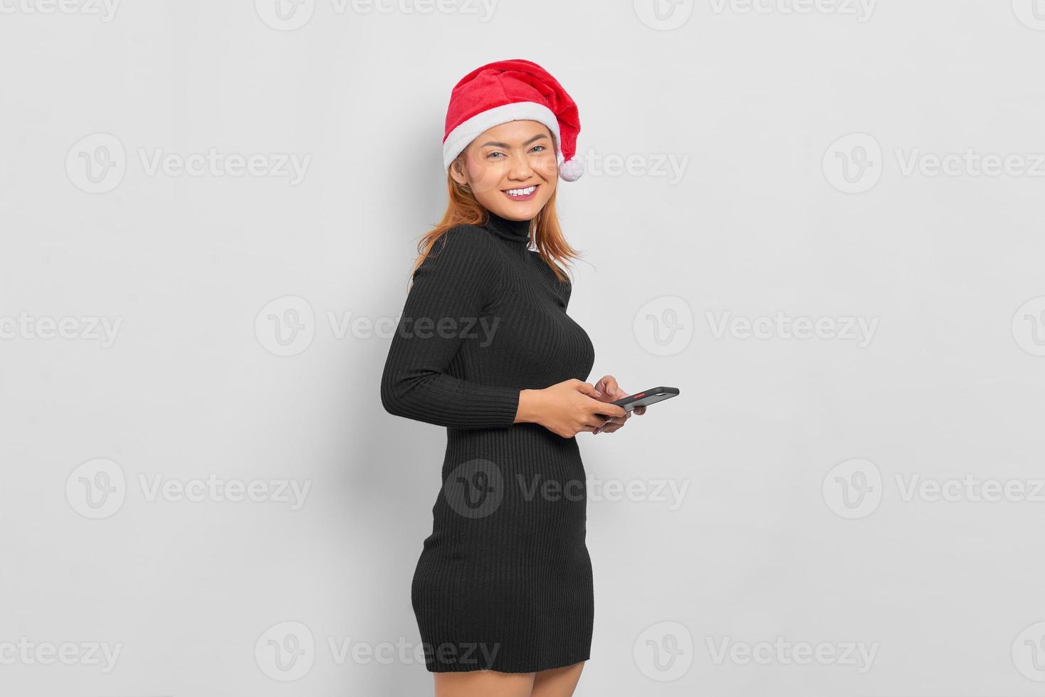 Portrait of smiling young Asian woman in Santa Claus hat using mobile phone isolated over white background photo