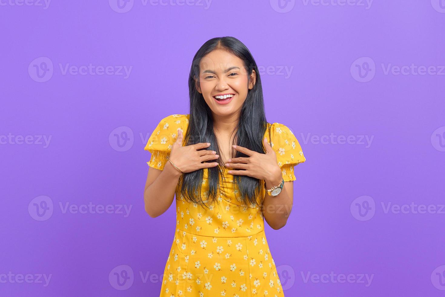 Cheerful young Asian woman put a hand on a chest and looking at camera on purple background photo