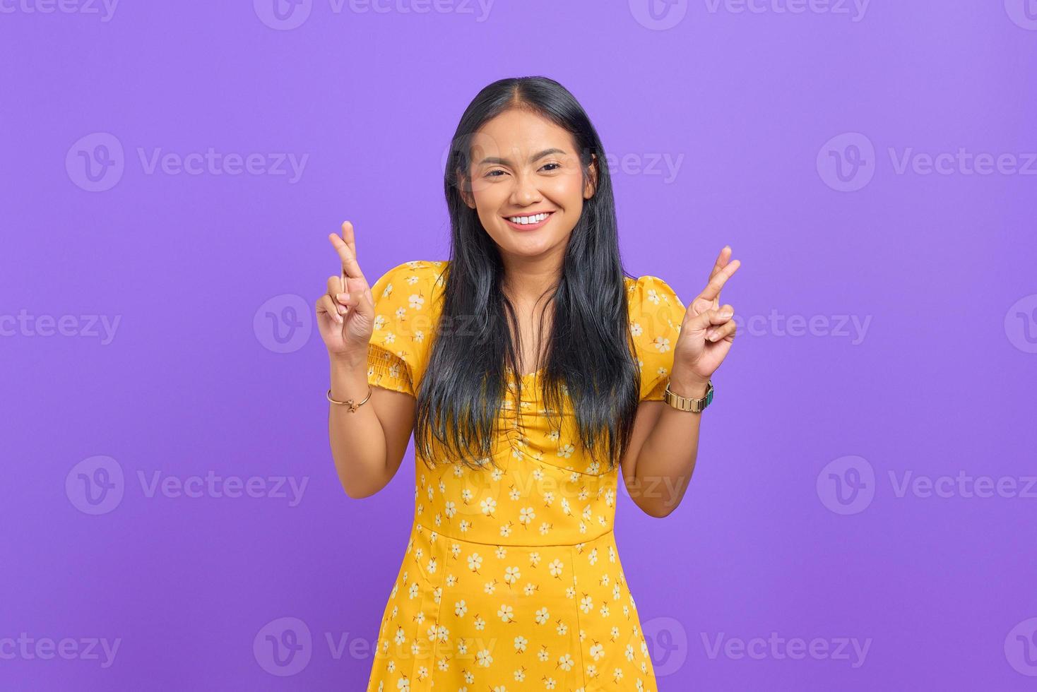 Sonriente joven mujer asiática pidiendo deseos con los dedos cruzados sobre fondo púrpura foto