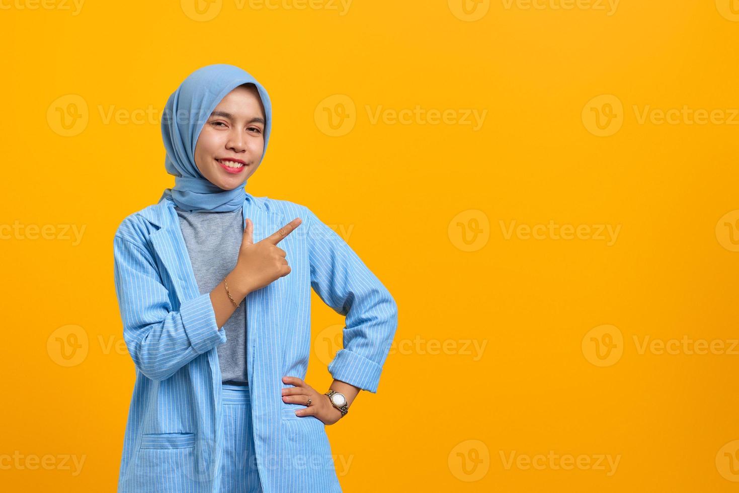 Cheerful young Asian woman pointing fingers aside at copy space isolated over yellow background photo