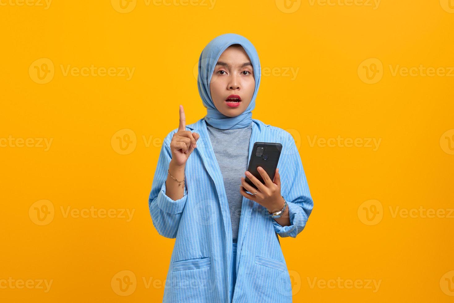 Surprised young Asian woman holding mobile phone and pointing finger up on yellow background photo