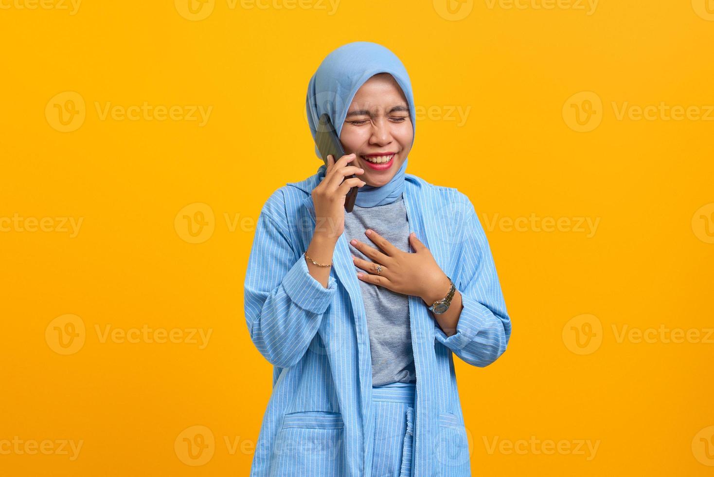 Cheerful young Asian woman talking on mobile phone and keeping palms on chest over yellow background photo