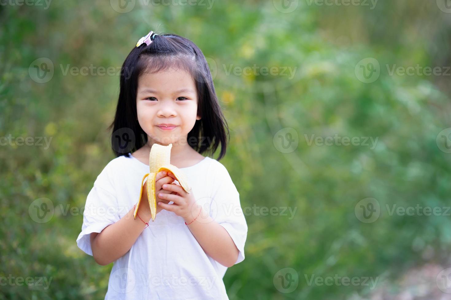 Asian girl sweet smile. Kid holding yellow banana in her hands. Child eating fruit. Copy space nature background. photo