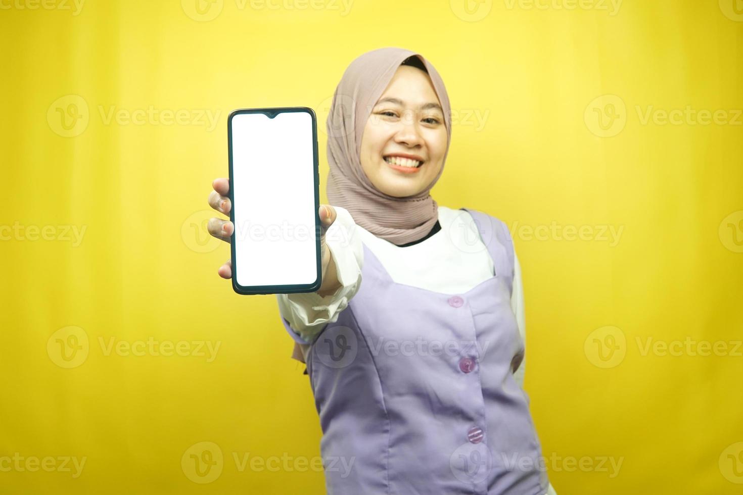 Hermosa joven musulmana asiática sonriendo con confianza y entusiasmo con las manos sosteniendo el teléfono inteligente, promoviendo la aplicación, mano de signo ok, buen trabajo, éxito, aislado sobre fondo amarillo foto