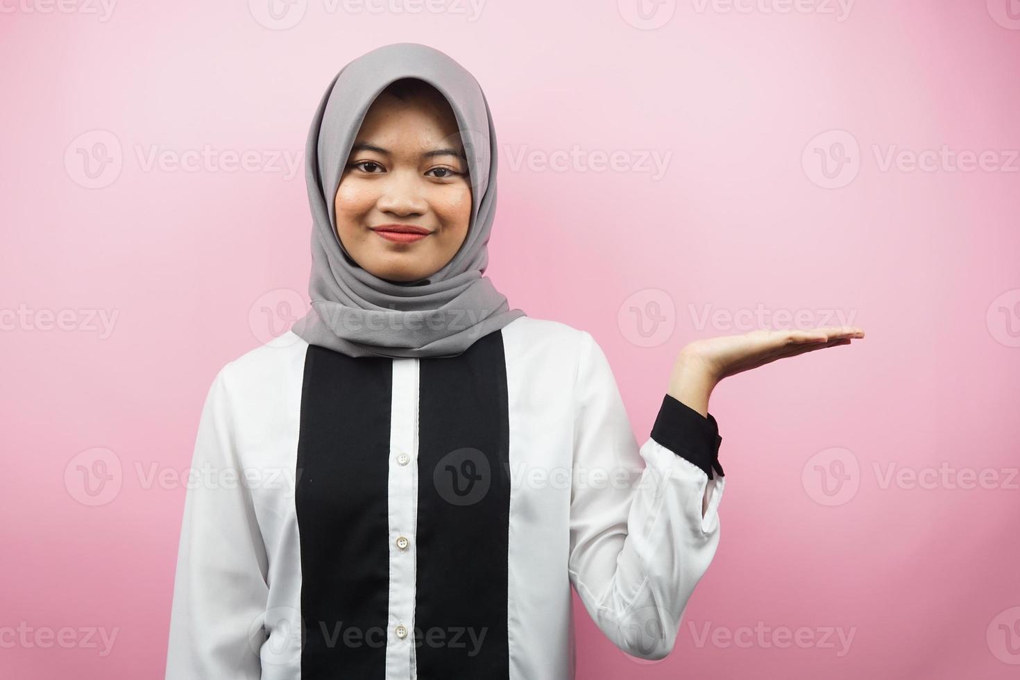 Beautiful young asian muslim woman with hands showing and presenting something in the direction of empty space, smiling confidently, excitedly, facing the camera, isolated on pink background photo