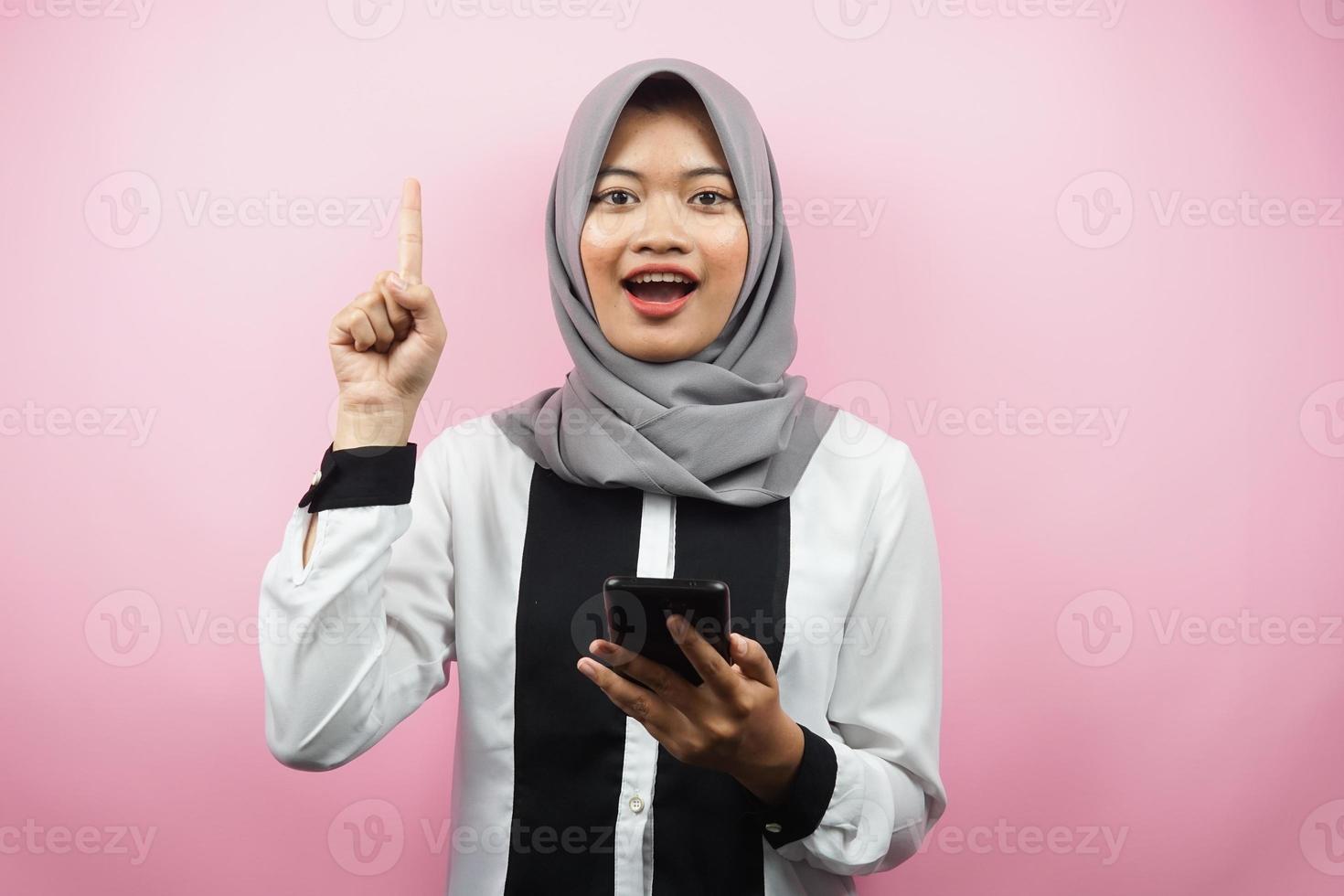 Beautiful young asian muslim woman smiling, shocked, surprised, getting an idea, with hands holding smartphone, isolated on pink background, advertising concept photo