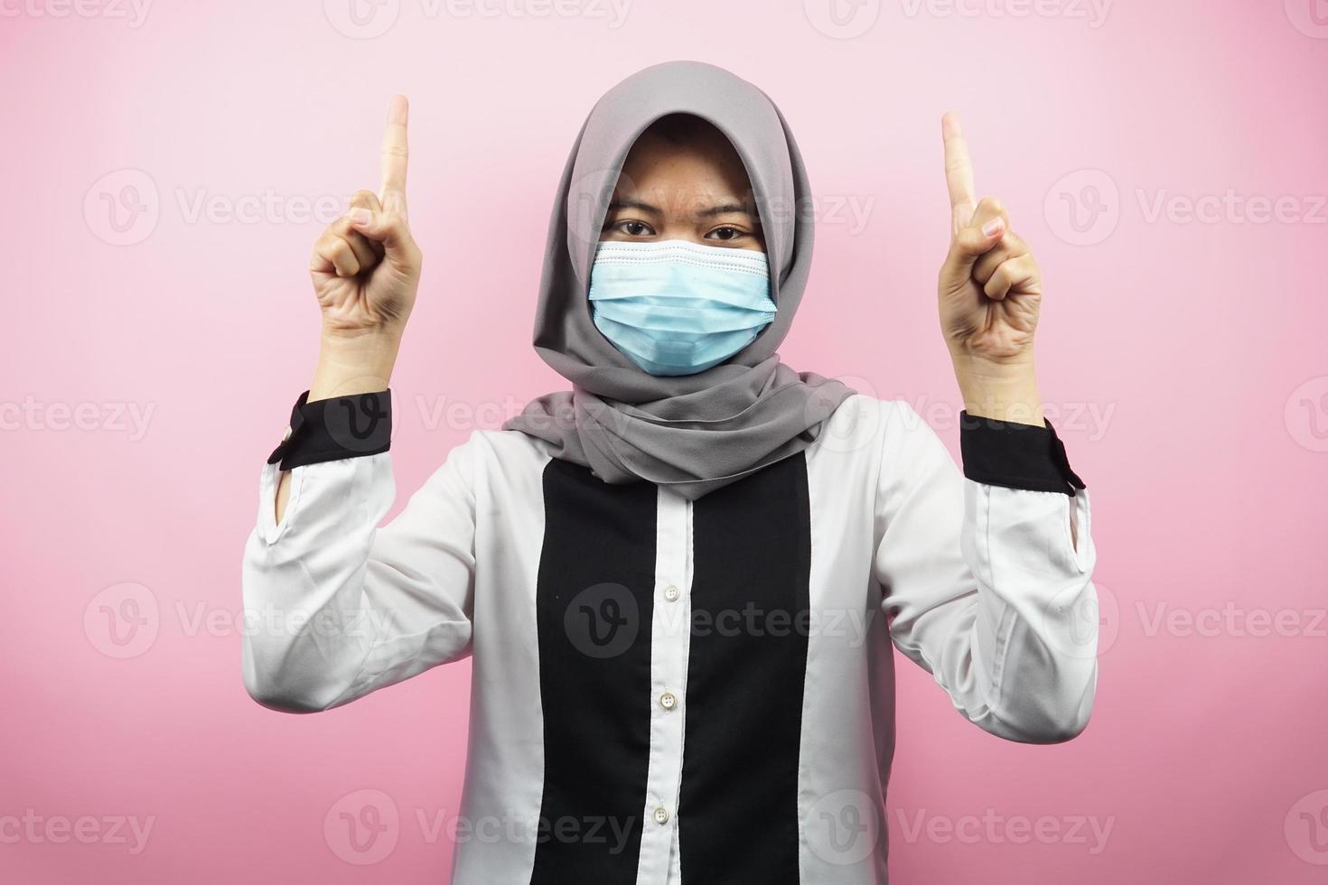 Mujer musulmana con máscara médica, mano apuntando al espacio vacío, mano apuntando hacia arriba presentando algo, aislado sobre fondo rosa foto