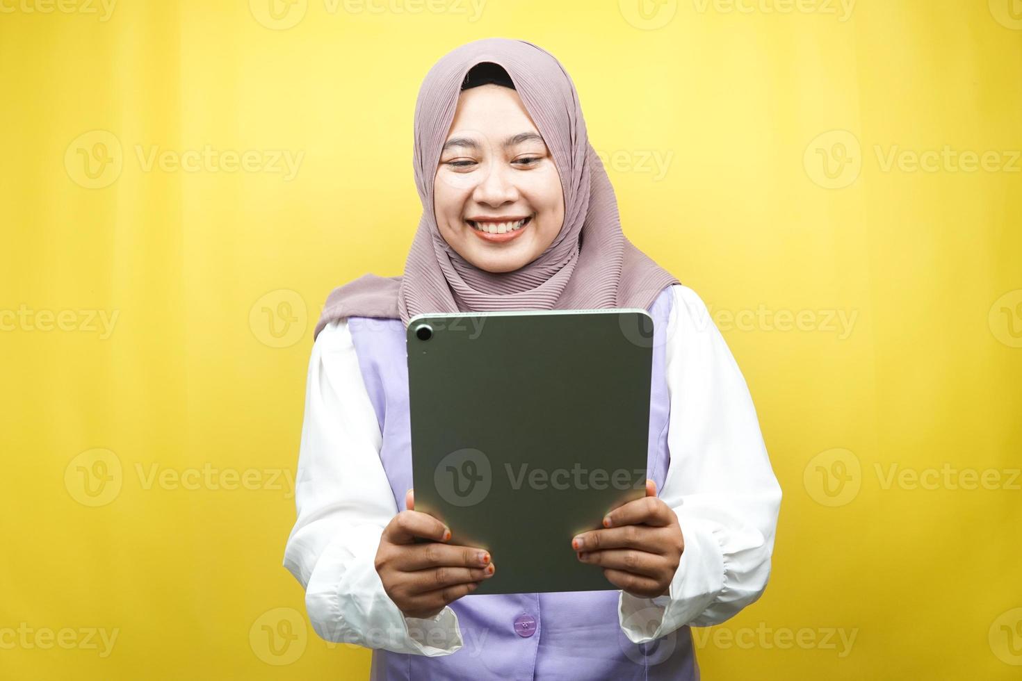 Beautiful young asian muslim woman smiling, excited and cheerful holding tablet, isolated on yellow background photo