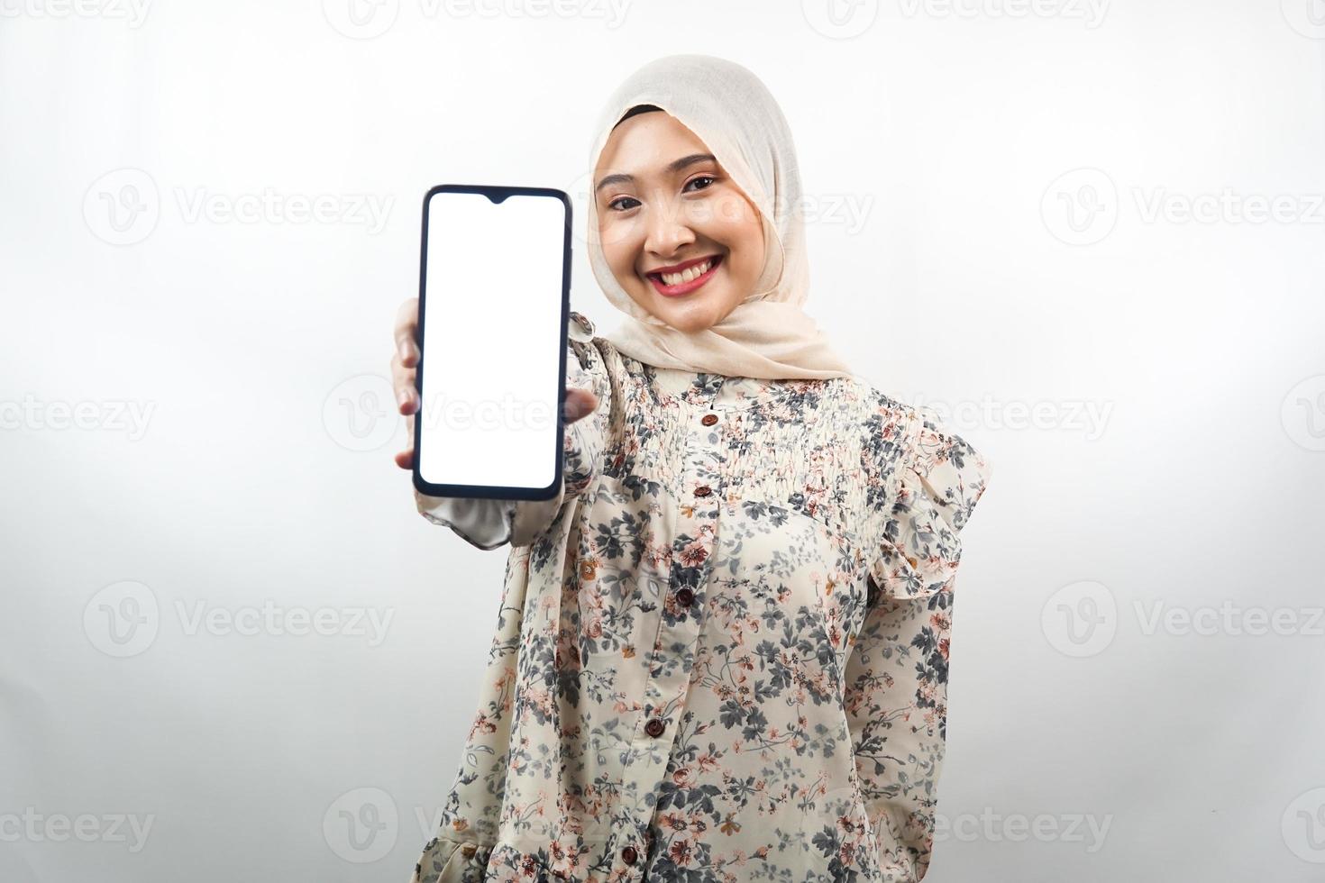 Hermosa joven musulmana asiática sonriendo confiada, entusiasta y alegre con las manos sosteniendo el teléfono inteligente, promoviendo la aplicación, promocionando algo, aislado sobre fondo blanco. foto