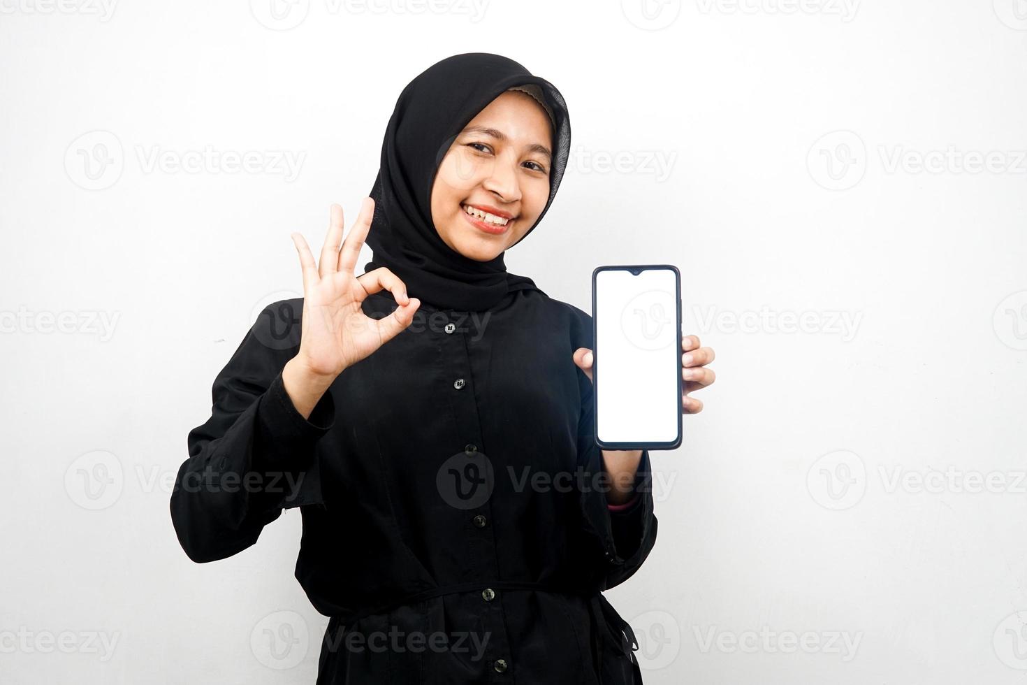 Beautiful young asian muslim woman with hands holding smartphone, promoting application, smiling confidently and excitedly, ok sign hand, good job, success, isolated on white background photo