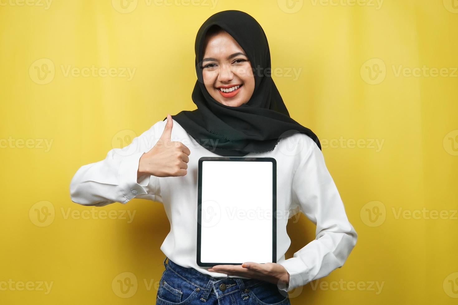 Beautiful young asian muslim woman smiling, excited and cheerful holding tablet with white or blank screen, promoting app, promoting product, presenting something, isolated on yellow background photo