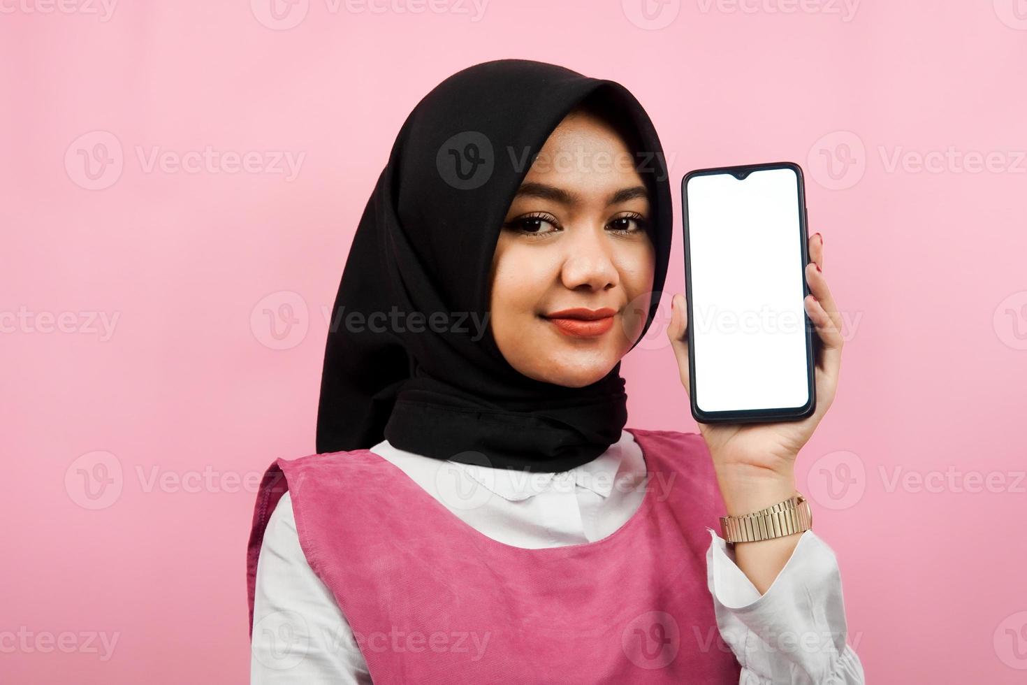 Close-up of beautiful and cheerful young Muslim woman holding smartphone with white or blank screen, promoting app, promoting something, isolated, advertising concept photo