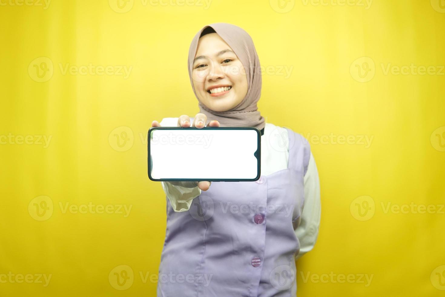 Beautiful young asian muslim woman smiling confident, enthusiastic and cheerful with hands holding smartphone, promoting application, promoting game, isolated on yellow background, advertising concept photo