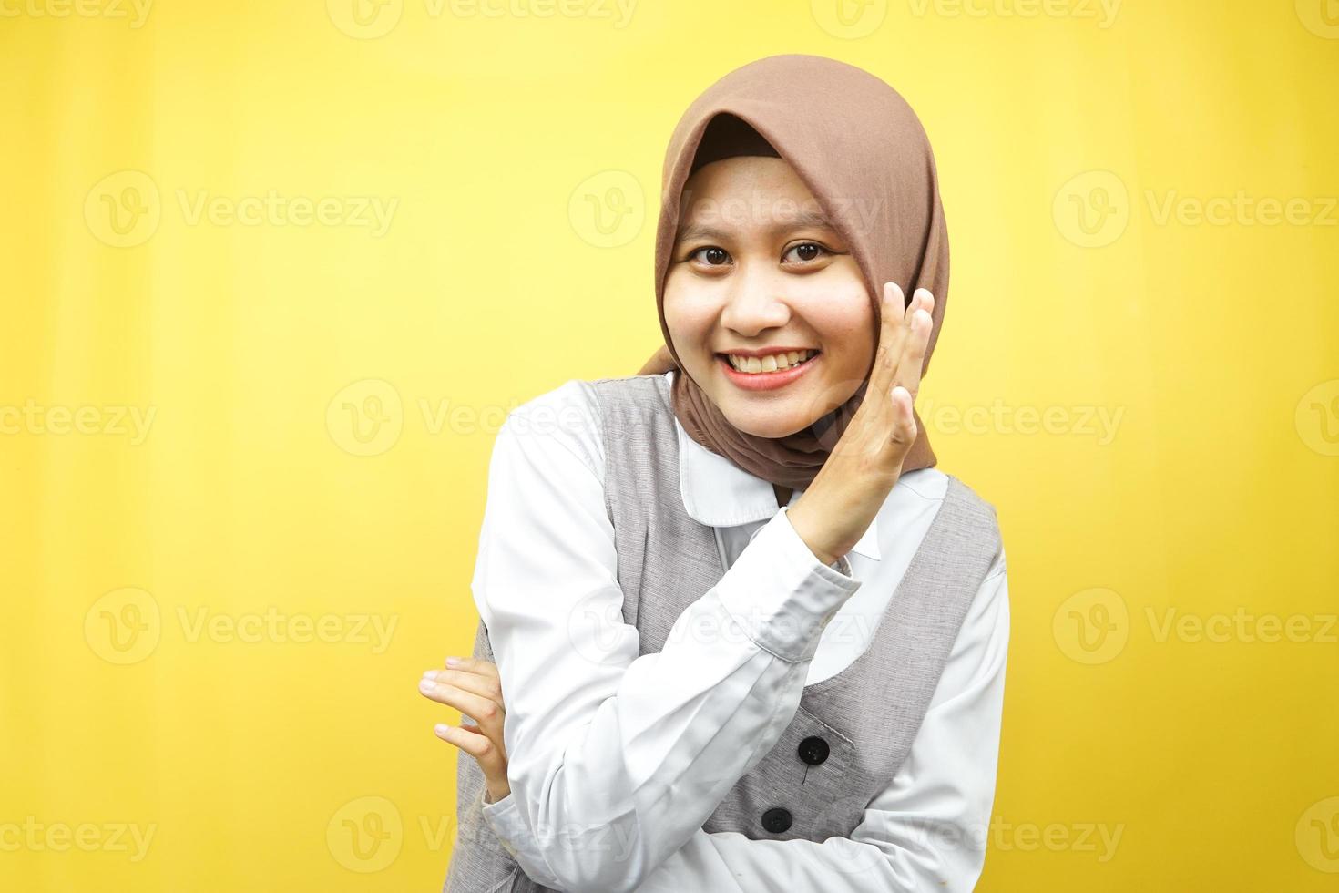 Beautiful young asian muslim woman smiling confidently and excitedly close to camera, whispering, telling secrets, speaking quietly, silent, isolated on yellow background photo