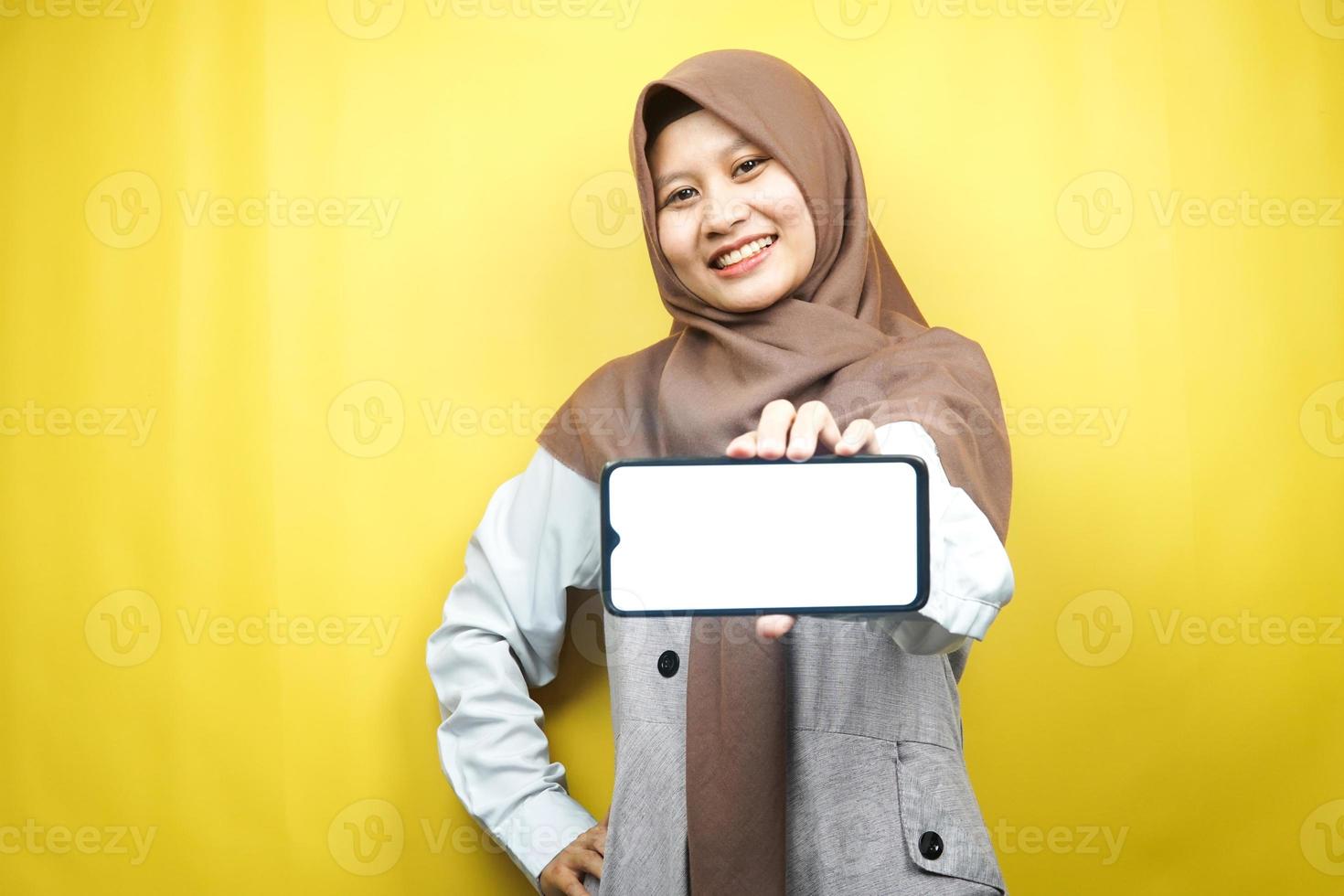 Beautiful young asian muslim woman smiling confident, enthusiastic and cheerful with hands holding smartphone, promoting application, promoting game, isolated on yellow background, advertising concept photo