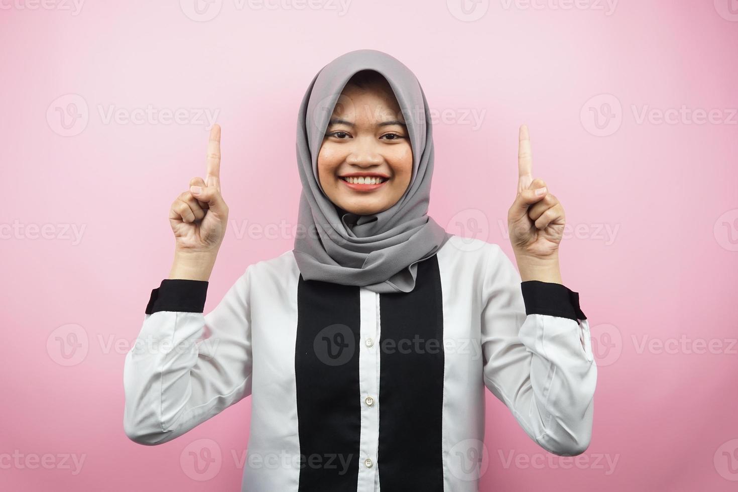 Beautiful young asian muslim woman smiling confident, enthusiastic and cheerful with hands pointing up presenting something, looking at camera isolated on pink background, advertising concept photo