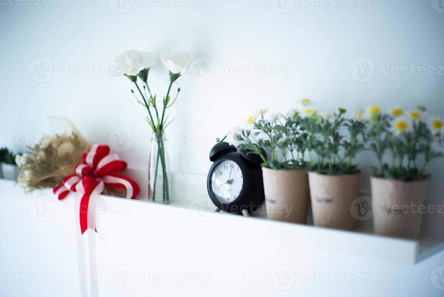 Vintage reloj despertador pone en el estante de madera blanca decorada por las flores en flor en la cerámica con fondo borroso de jarrón foto