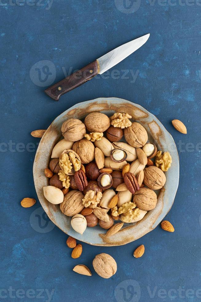 mezcla de nueces en un plato - nuez, almendras, nueces, macadamia. comida vegana saludable. alimentación limpia, dieta equilibrada. mesa azul brillante, cuchillo para abrir concha. vista superior, vertical foto