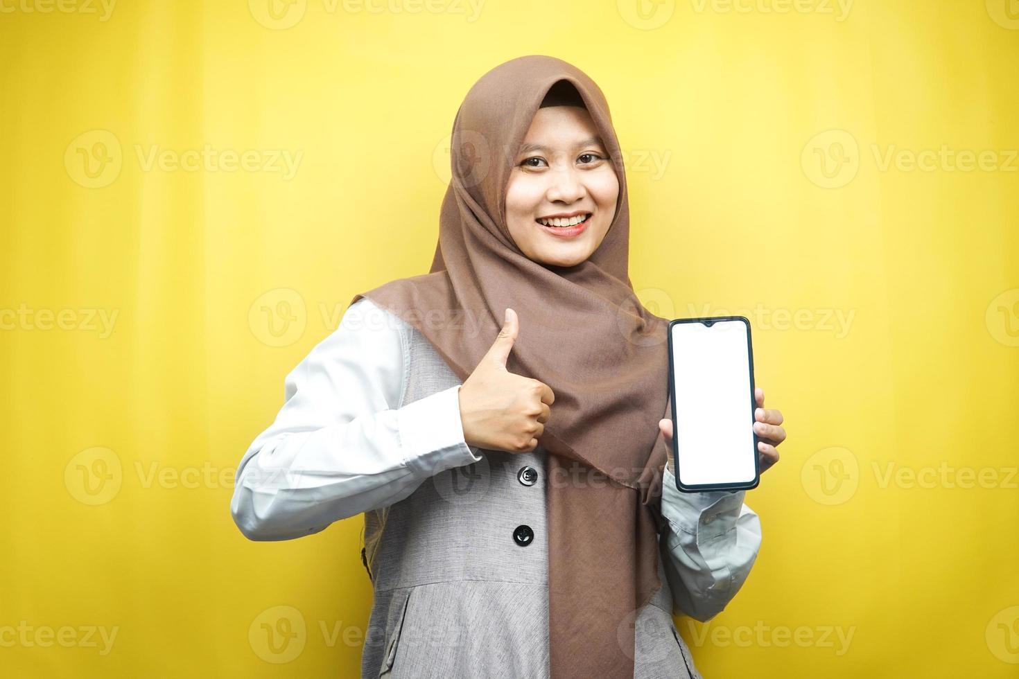 Hermosa joven musulmana asiática sonriendo confiada, entusiasta y alegre con la mano que sostiene el teléfono inteligente, promocionando algo, la aplicación de promoción de la mano, aislada sobre fondo amarillo foto