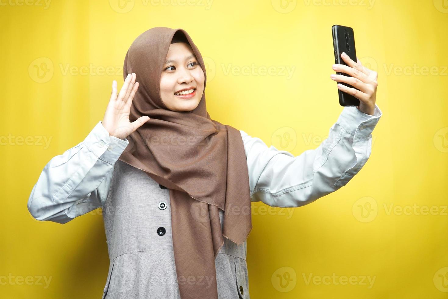 Beautiful young asian muslim woman smiling confident, enthusiastic and cheerful with hands holding smartphone, calling someone, making video call, taking selfie, isolated on yellow background photo