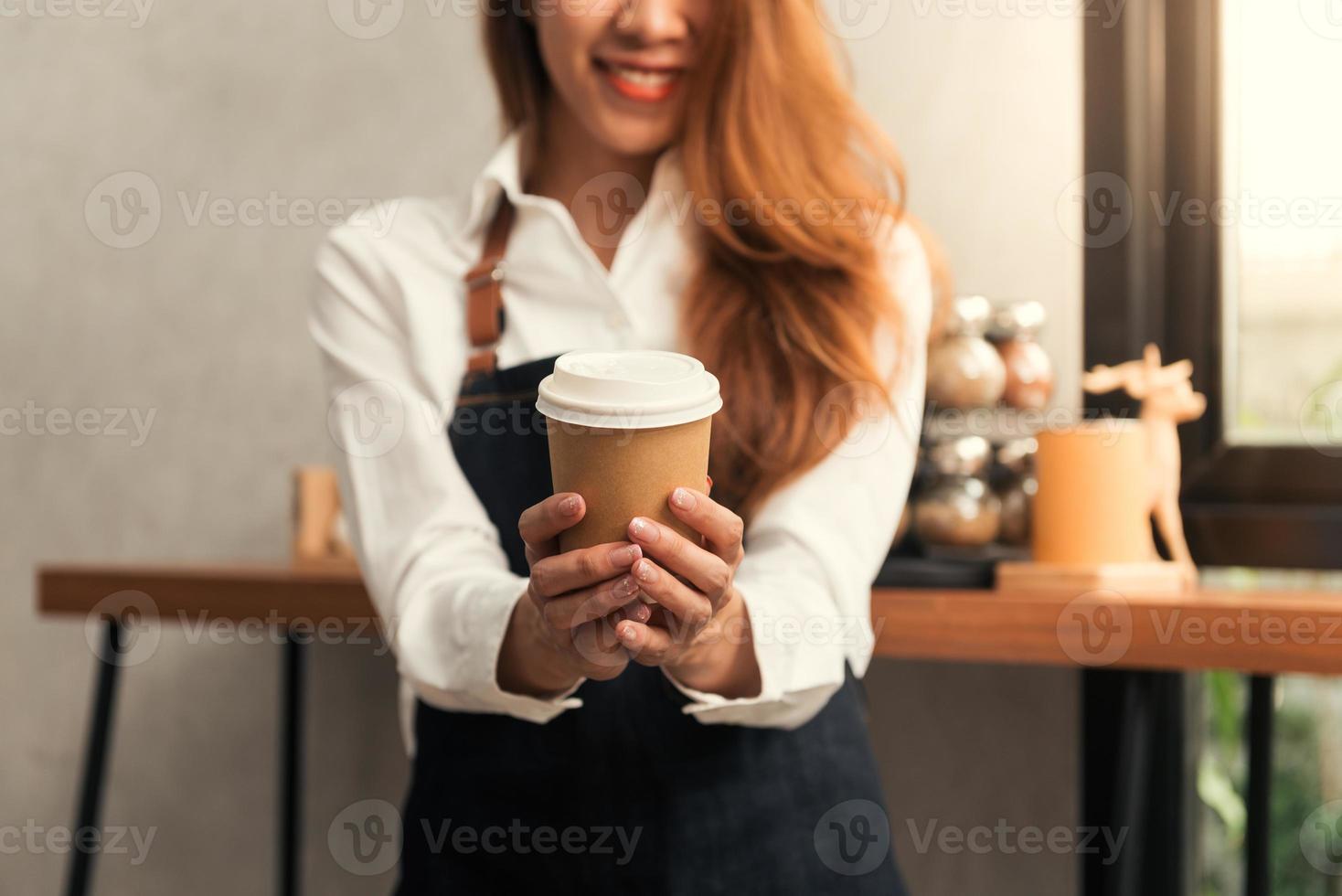 Close up of a young Asian female barista hold a cup of coffee serving to her customer with smile surrounded with bar counter background. Young female barista and her small shop. Food and drink concept photo