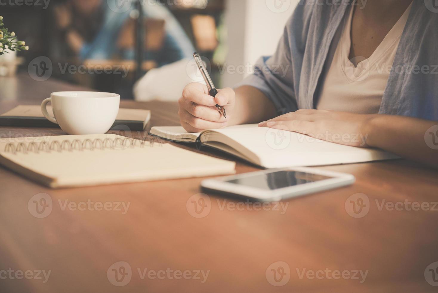 Young business woman in white dress sitting at table in cafe and writing in notebook. Asian woman talking smartphone and cup of coffee. Freelancer working in coffee shop. photo