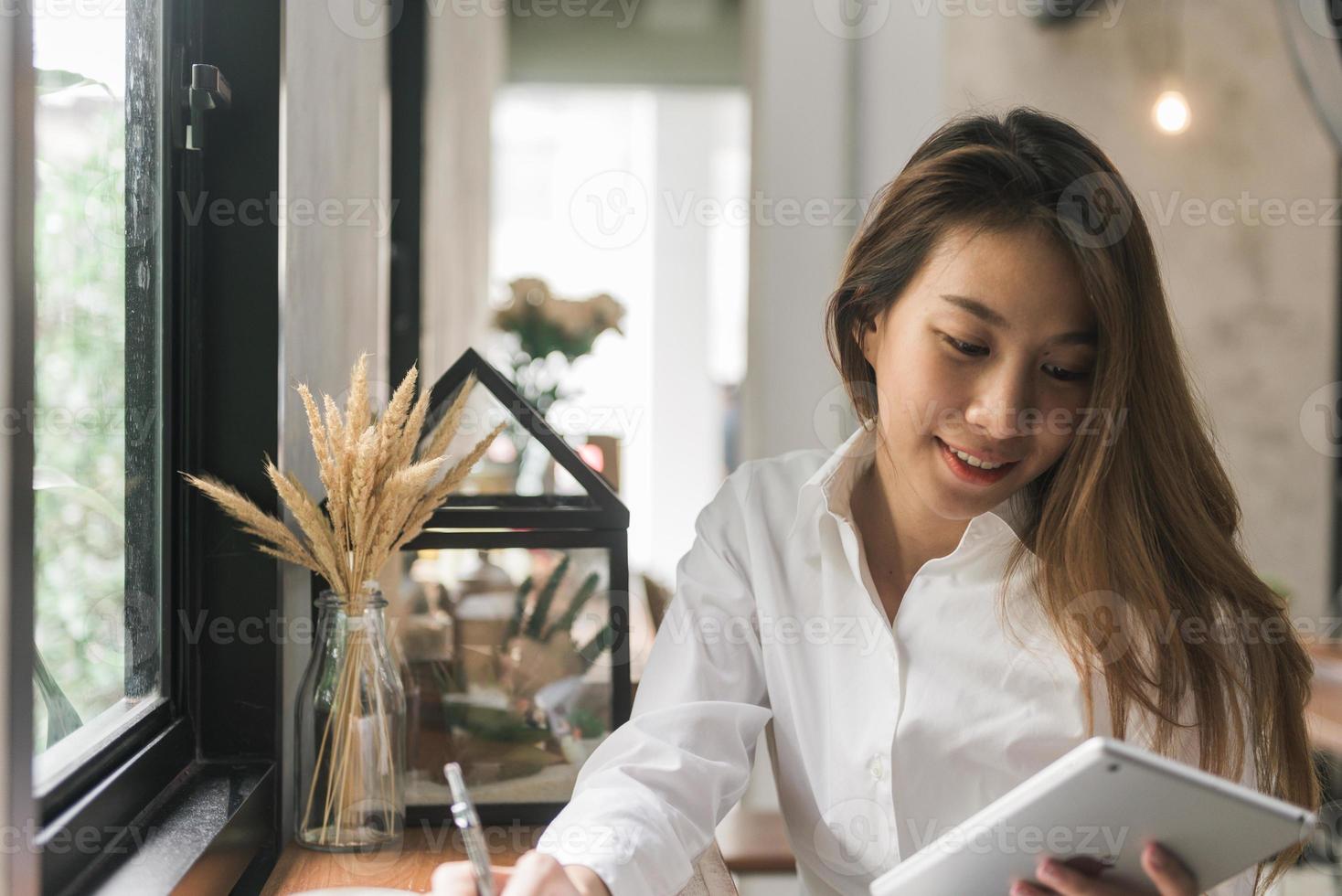 mujer de negocios joven en vestido blanco sentado a la mesa en la cafetería y escribiendo en el cuaderno. mujer asiática con tableta y taza de café. autónomo que trabaja en la cafetería. aprendizaje de los estudiantes en línea. foto