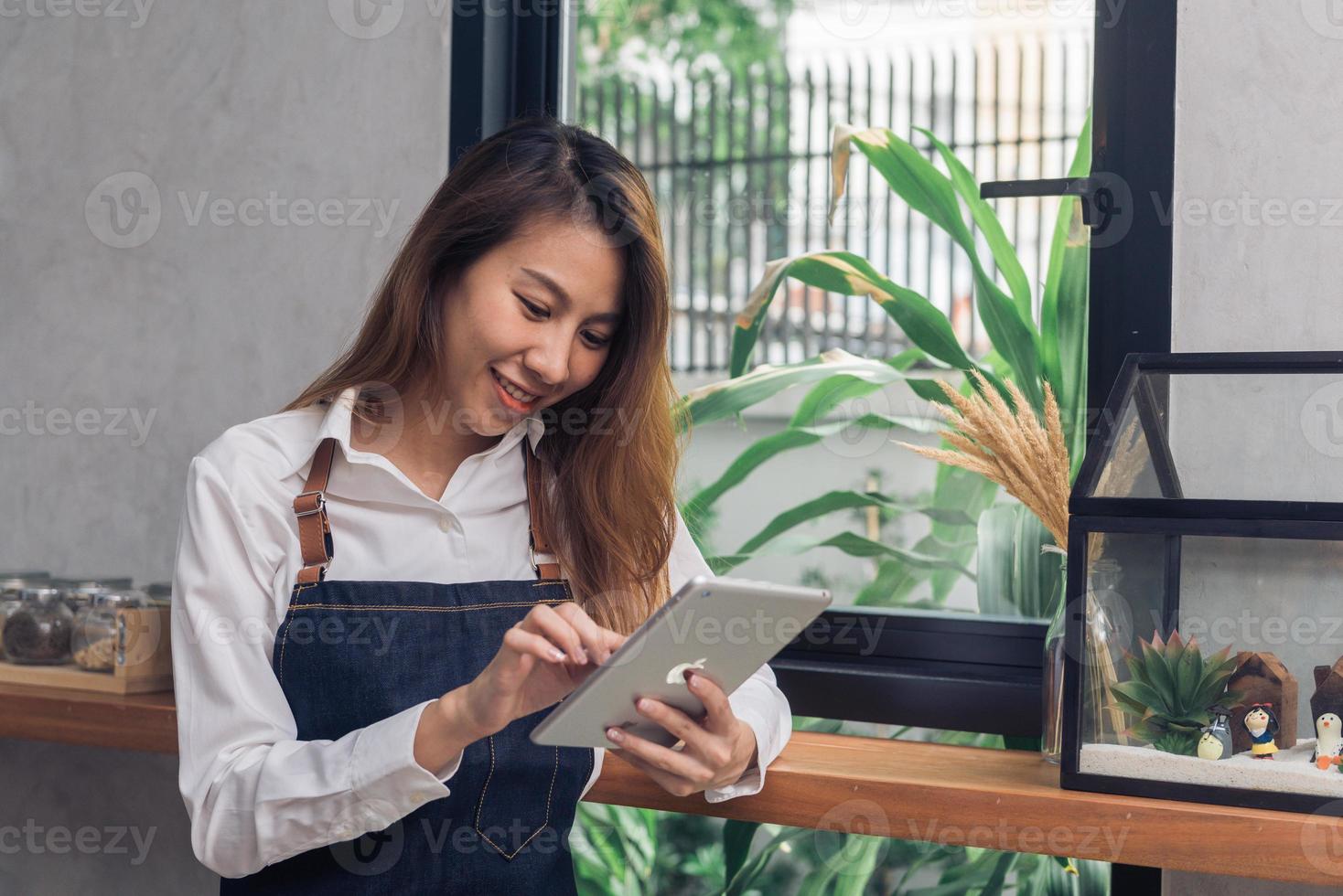 Young Asian female barista using electronic tablet in her own coffee shop while takeing break in warm afternoon. Young female barista and her modern small coffee shop. Food and drink industry concept. photo