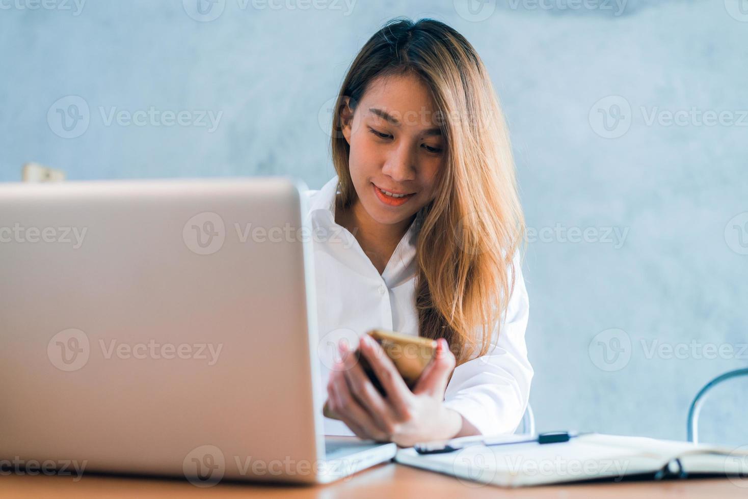 mujer asiática joven que trabaja con la computadora portátil en un escritorio con su sonrisa. joven asiática trabajando el fin de semana con su computadora portátil en un cálido día de luz solar. portátil trabajando en el concepto de casa. foto