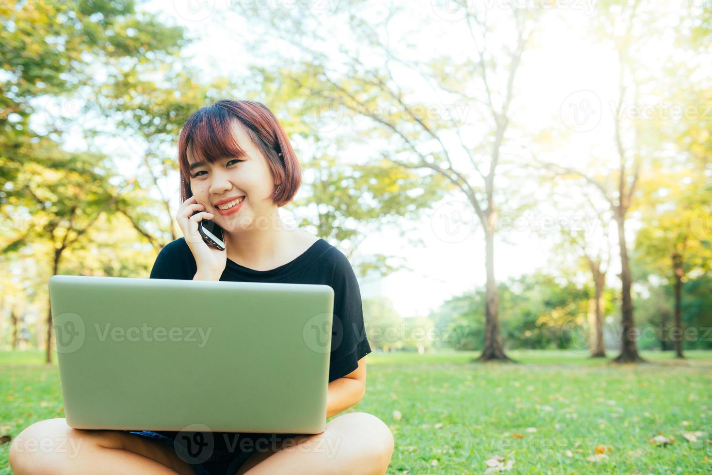 Linda mujer asiática sonríe y habla por teléfono móvil mientras está sentado en el parque día de primavera. mujer asiática que usa el teléfono inteligente y la computadora portátil con sensación de relajación y cara sonriente. conceptos de estilo de vida y tecnología. foto