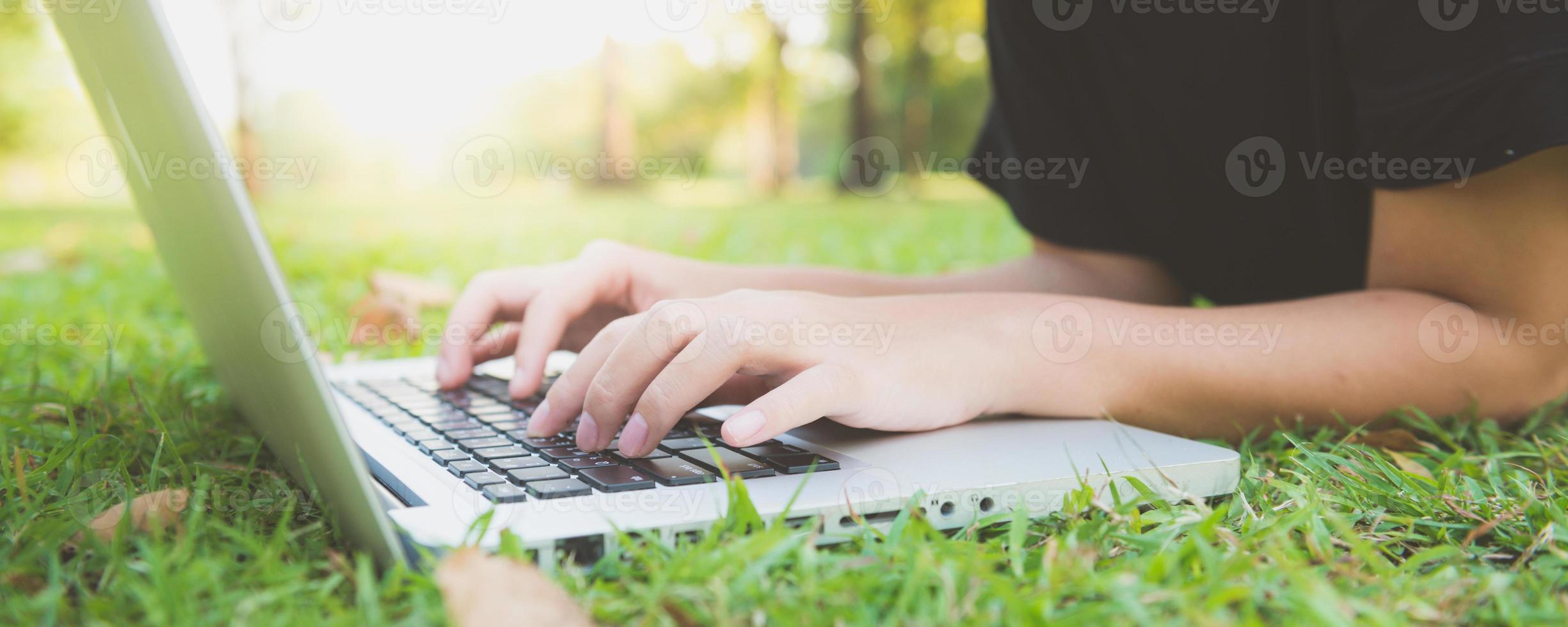Young asian woman's legs on the green grass with open laptop. Girl's hands on keyboard. Distance learning concept. Happy hipster young asian woman working on laptop in park. Panoramic banner. photo