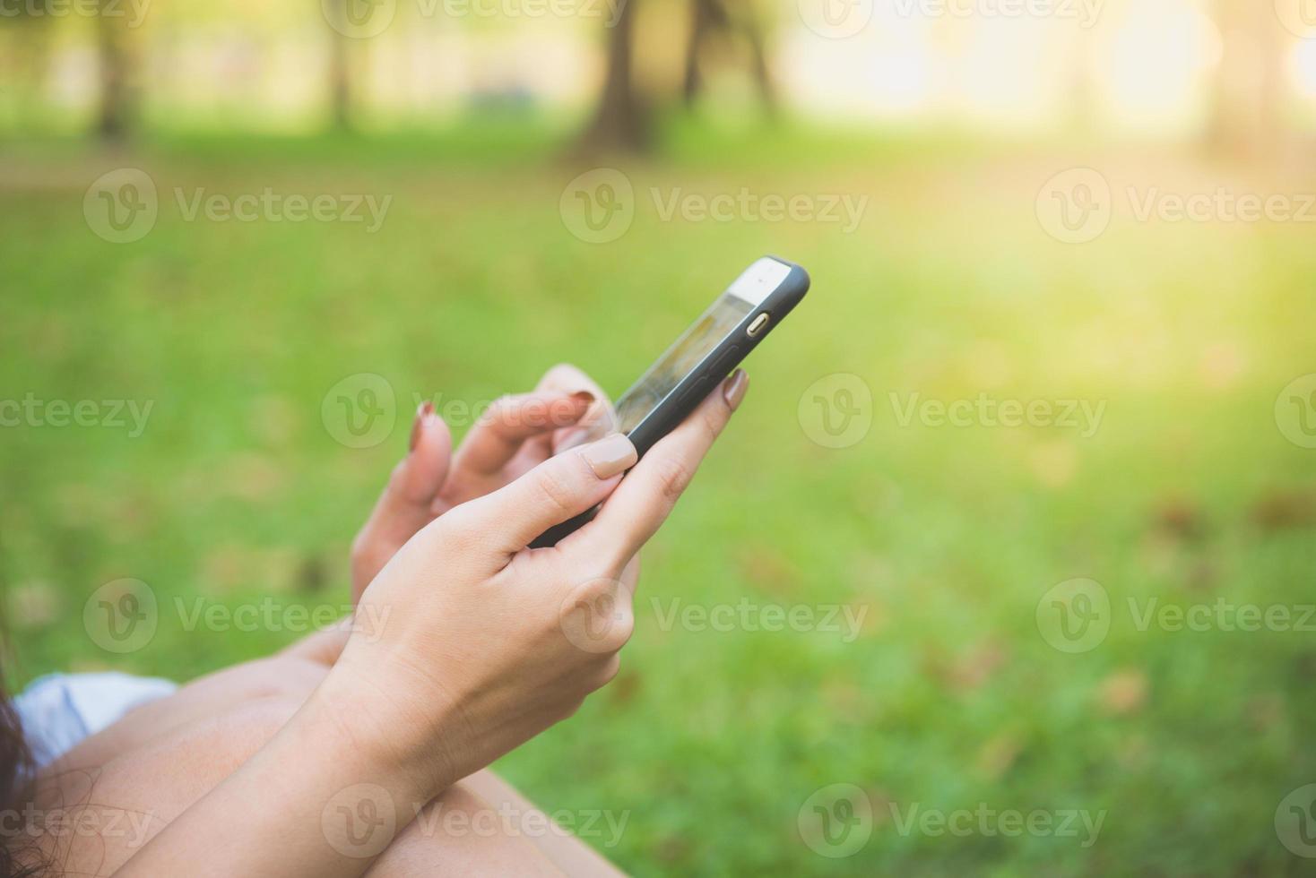 Linda mujer asiática leyendo un agradable mensaje de texto en el teléfono móvil mientras está sentado en el parque el día de la primavera. mujer asiática que usa el teléfono inteligente con sensación de relajación y cara sonriente. conceptos de estilo de vida y tecnología. foto