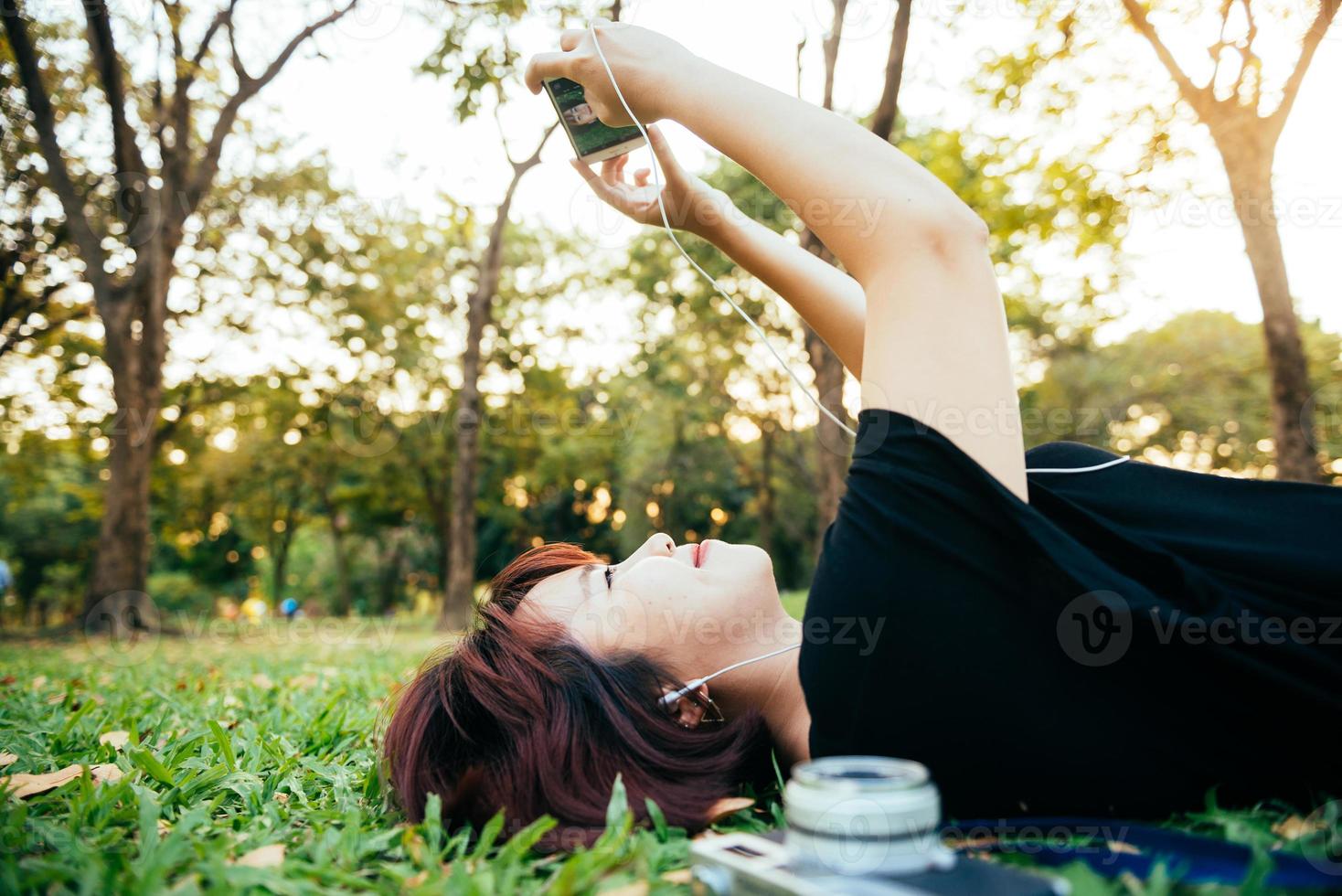 joven asiática tendida en la hierba verde escuchando música en el parque con una emoción escalofriante. mujer joven relajándose en la hierba con su cámara al lado. actividad al aire libre en el concepto de parque. foto