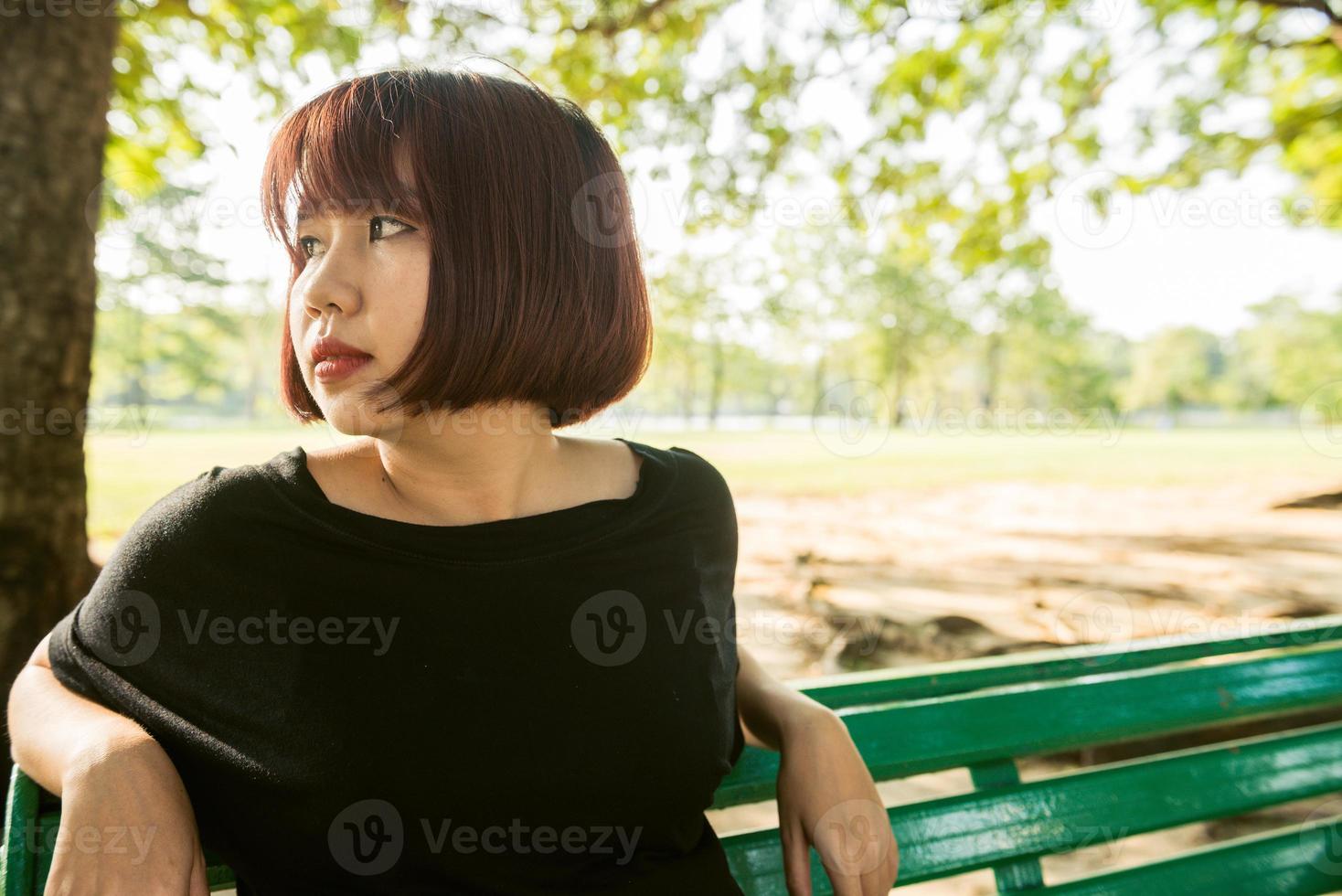 Young Asian woman sitting outdoor on the public bench and lying along the bench in the park in warm sunlight day. Young Woman relax her mind and body in the park. Outdoor activity in the park concept. photo