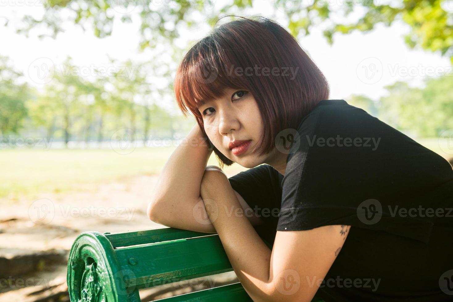 joven asiática sentada al aire libre en el banco público y acostada a lo largo del banco en el parque en un cálido día de luz solar. mujer joven relajar su mente y cuerpo en el parque. actividad al aire libre en el concepto de parque. foto