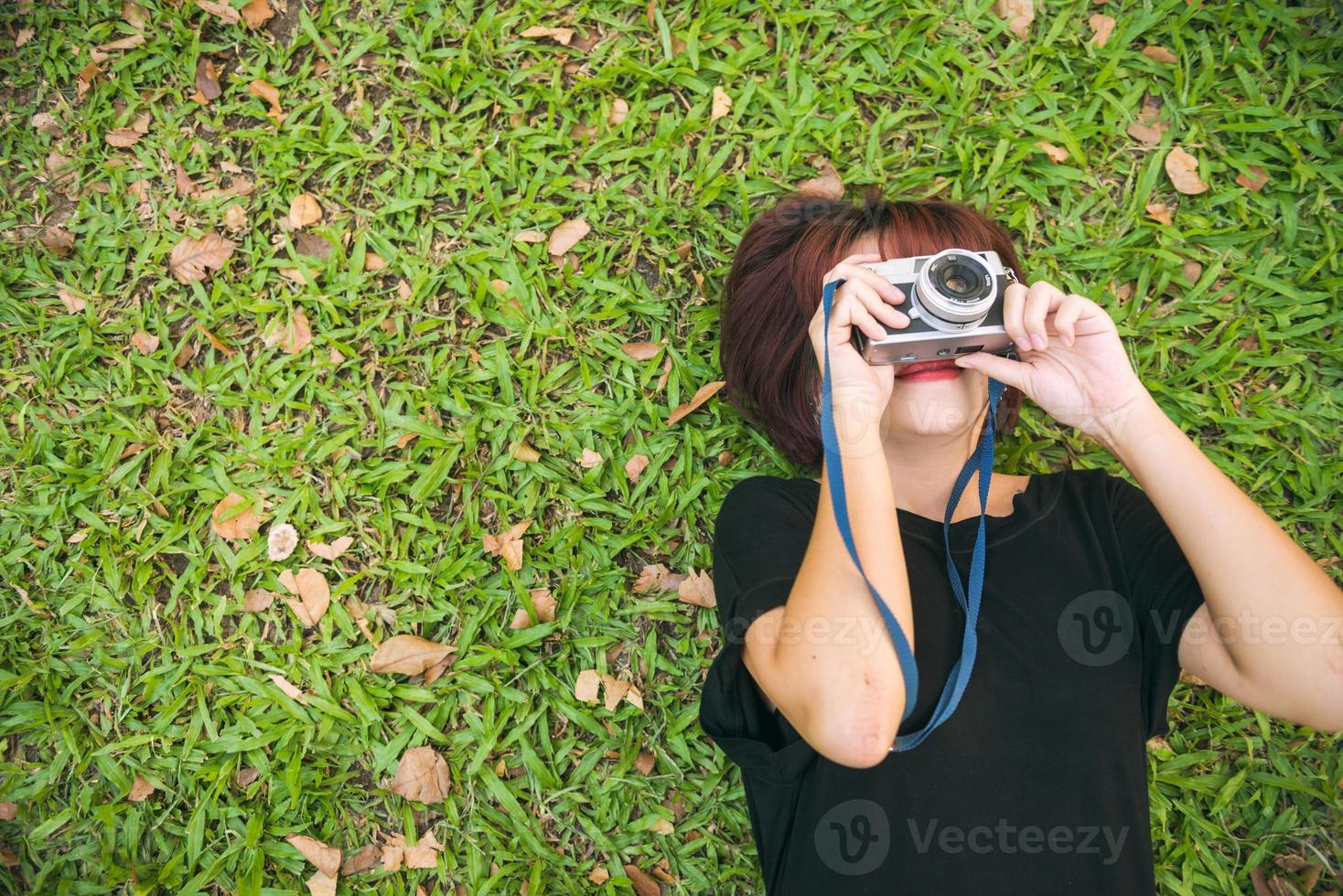Close up of a young Asian woman laying on the green grass chilly and shoot a photo with her camera. Young woman relaxing on the grass with her camera beside. Outdoor activity in the park concept.