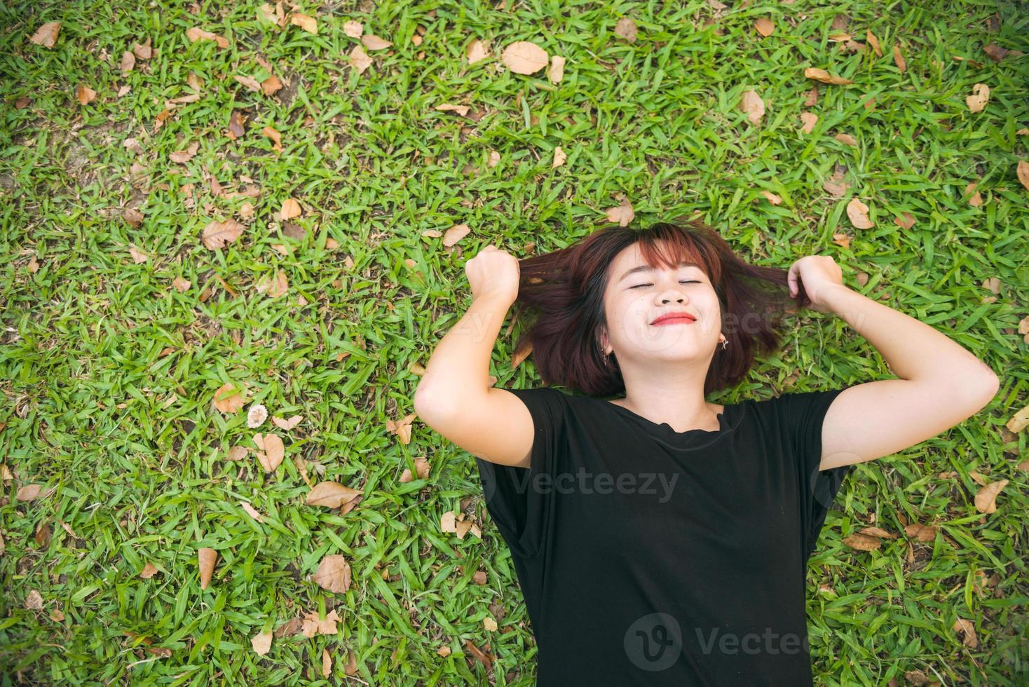 Young Asian woman laying on the green grass listening to music in the park with a chill emotion. Young woman relaxing on the grass with her music playlist. Outdoor activity in the park concept. photo