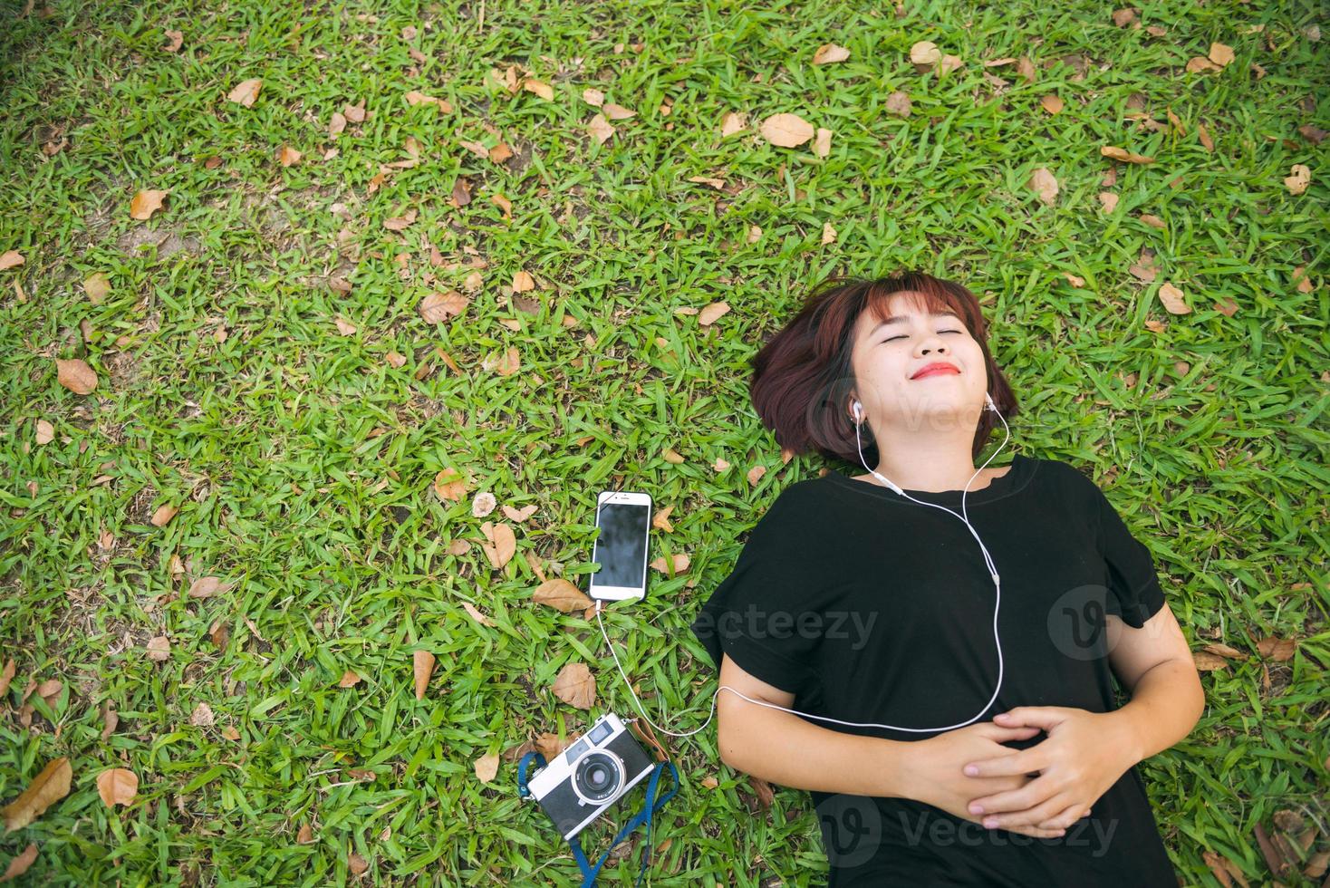 joven asiática tendida en la hierba verde escuchando música en el parque con una emoción escalofriante. mujer joven relajándose en el césped con su lista de reproducción de música. actividad al aire libre en el concepto de parque. foto