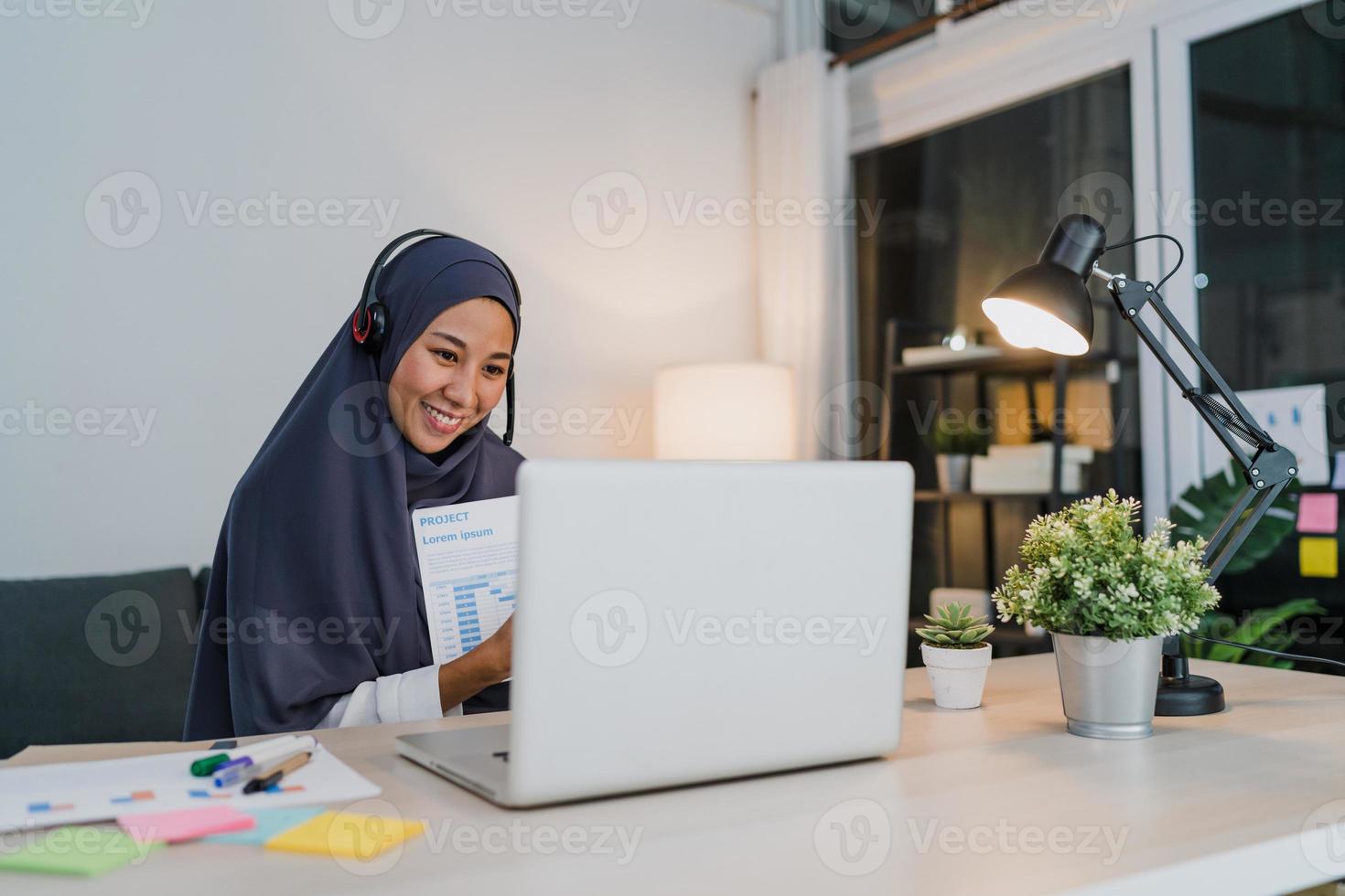 Asia muslim lady wear headphone using laptop talk to colleagues about sale report in conference video call while working from home office at night. Social distancing, quarantine for corona virus. photo
