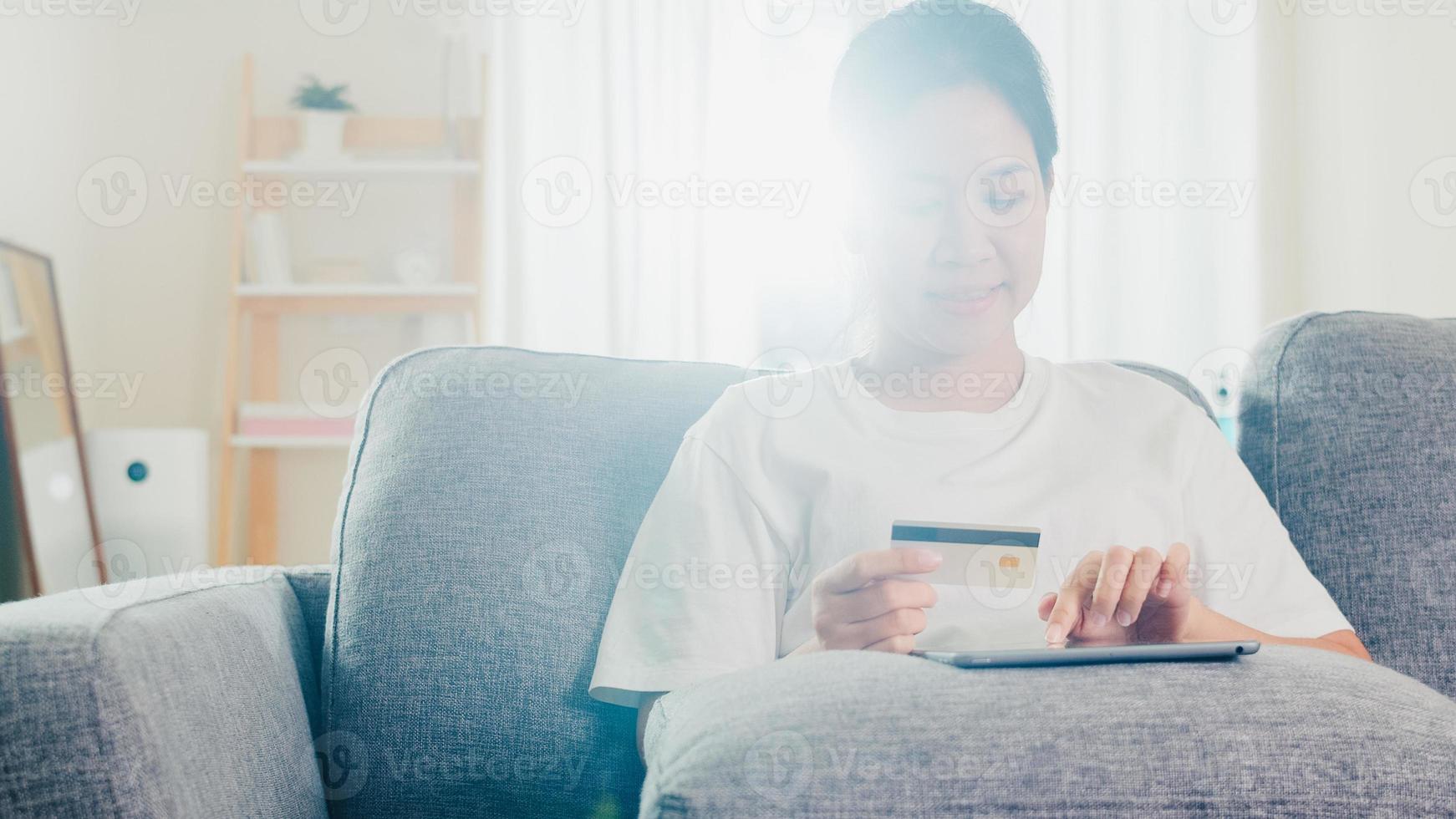 Mujer asiática que usa tableta, compra con tarjeta de crédito y compra Internet de comercio electrónico en la sala de estar de su casa cuando el distanciamiento social se queda en casa y el tiempo de auto cuarentena, compras en línea, concepto de coronavirus. foto