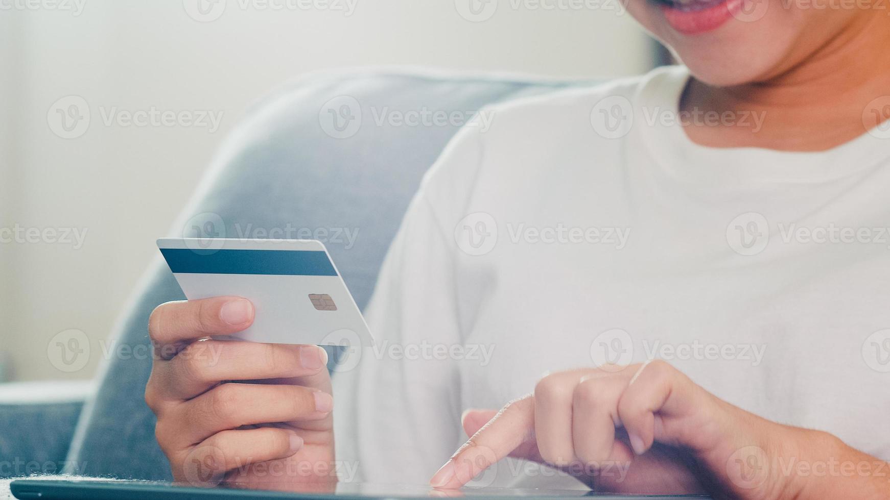 Asian woman using tablet, credit card buy and purchase e-commerce internet in living room from home when social distancing stay at home and self quarantine time, online shopping, coronavirus concept. photo