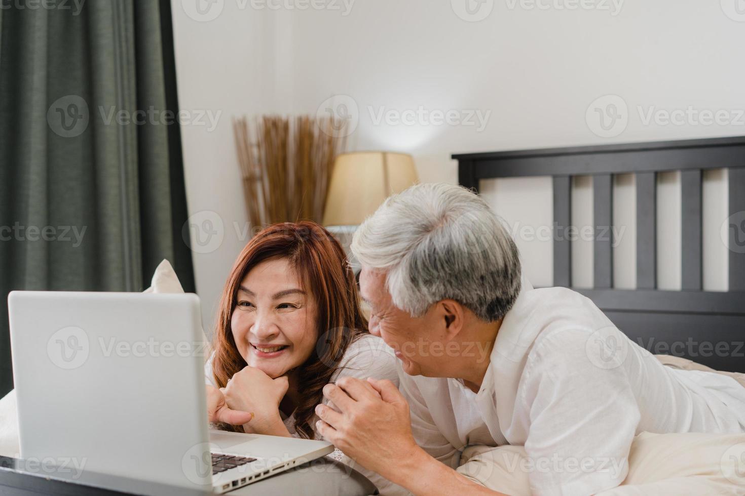 Asian senior couple using laptop at home. Asian Senior Chinese grandparents, husband and wife happy after wake up, watching movie lying on bed in bedroom at home in the morning concept. photo