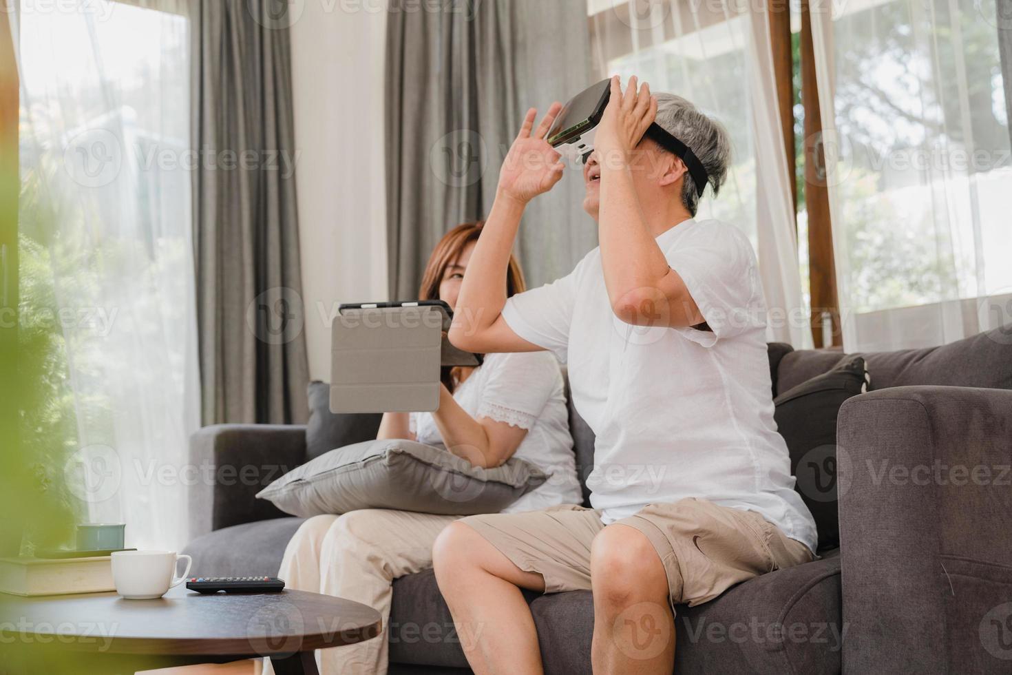 Asian elderly couple using tablet and virtual reality simulator playing games in living room, couple feeling happy using time together lying on sofa at home. Lifestyle Senior family at home concept. photo