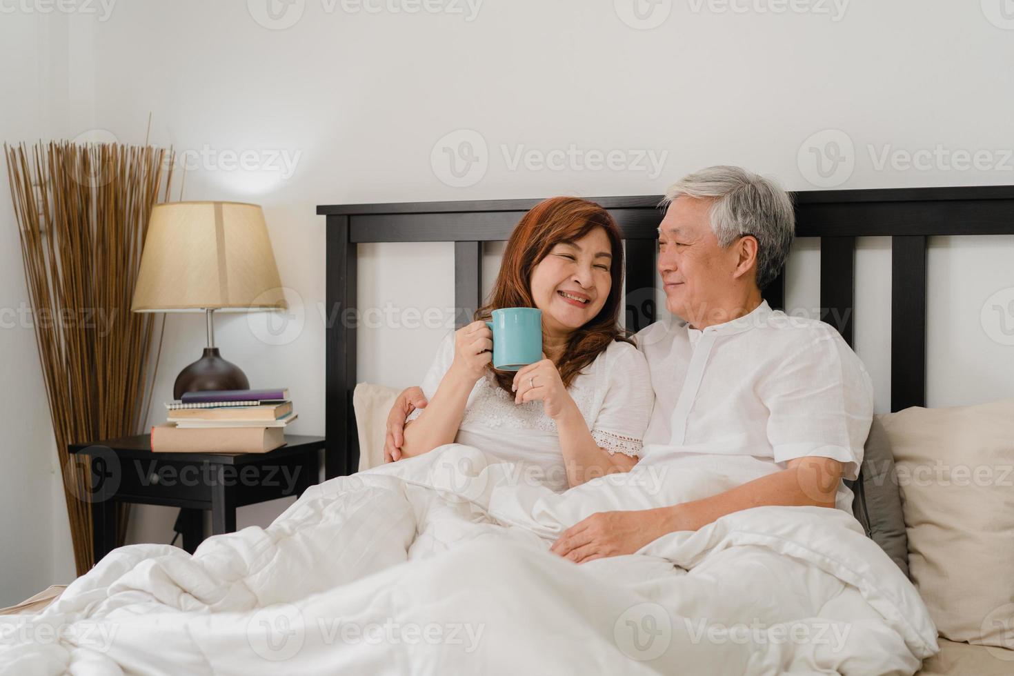 pareja senior asiática hablando en la cama en casa. Abuelos chinos mayores asiáticos, esposo y esposa felices beben café después de despertarse mientras está acostado en la cama en el dormitorio en casa en el concepto de la mañana. foto