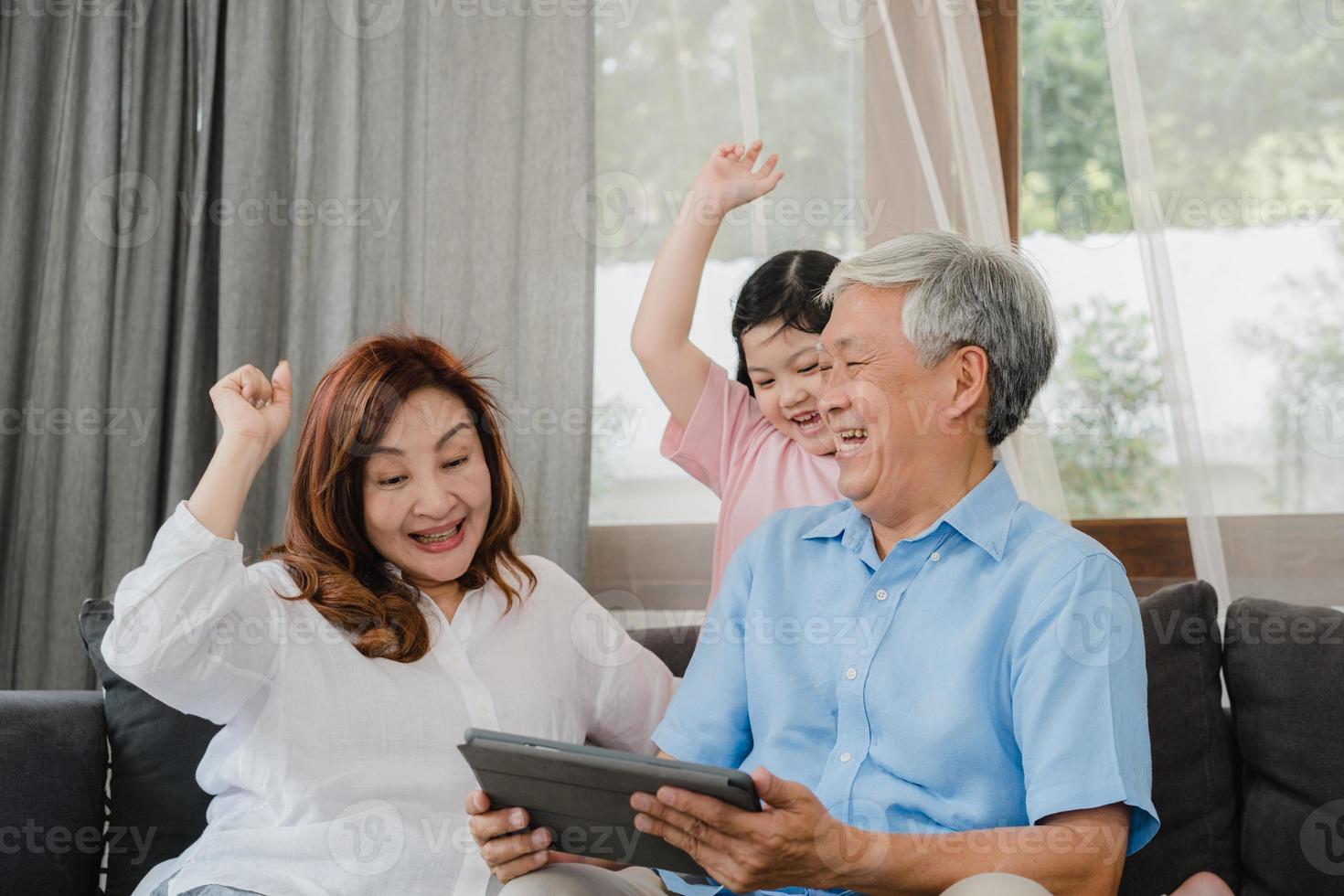 abuelos asiáticos y nieta usando tableta en casa. Senior chino, abuelo y abuela felices pasan tiempo en familia relajarse con una niña revisando las redes sociales, acostada en el sofá en el concepto de sala de estar foto